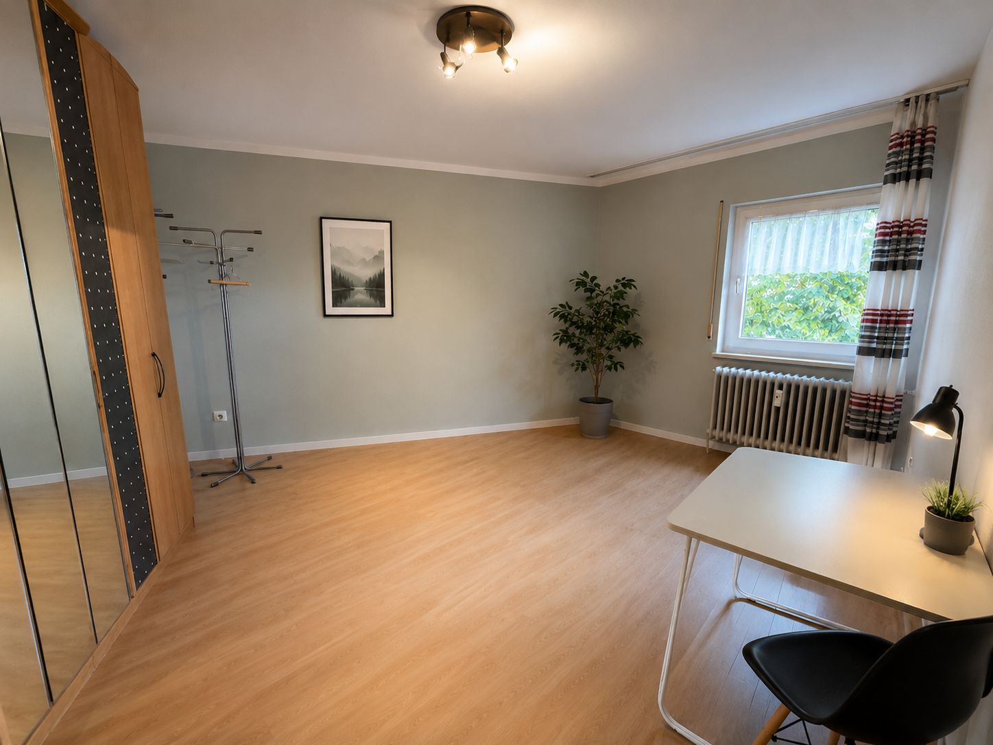 Helles Zimmer mit Schreibtisch, Stuhl, Kleiderschrank, Pflanze und Fenster mit Vorhängen.