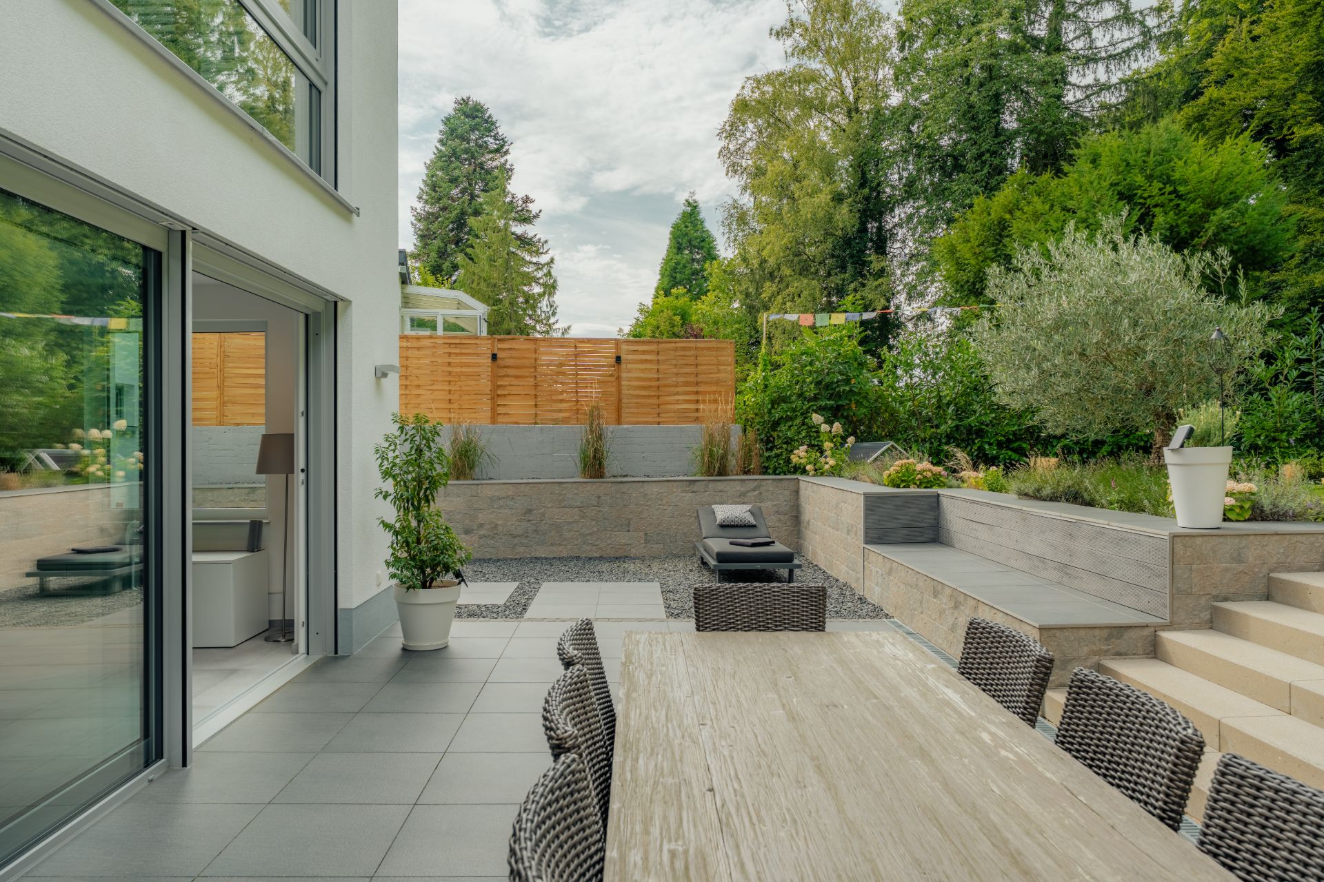 Moderne Terrasse mit großem Holztisch, Stühlen, Pflanzen und Garten im Hintergrund.