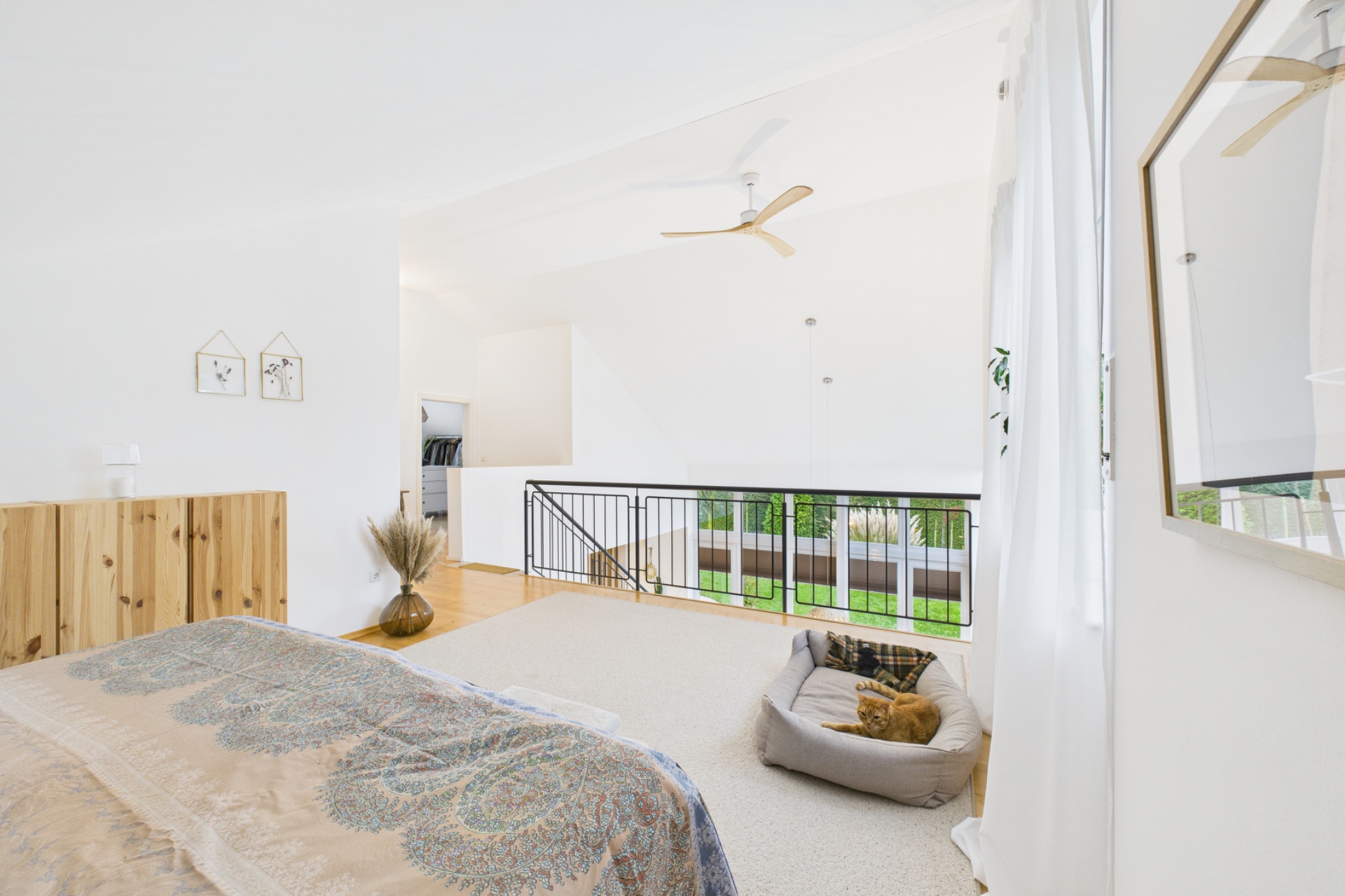Helles Schlafzimmer mit Bett, Holzkommode, Deckenventilator und Hundebett am Fenster.