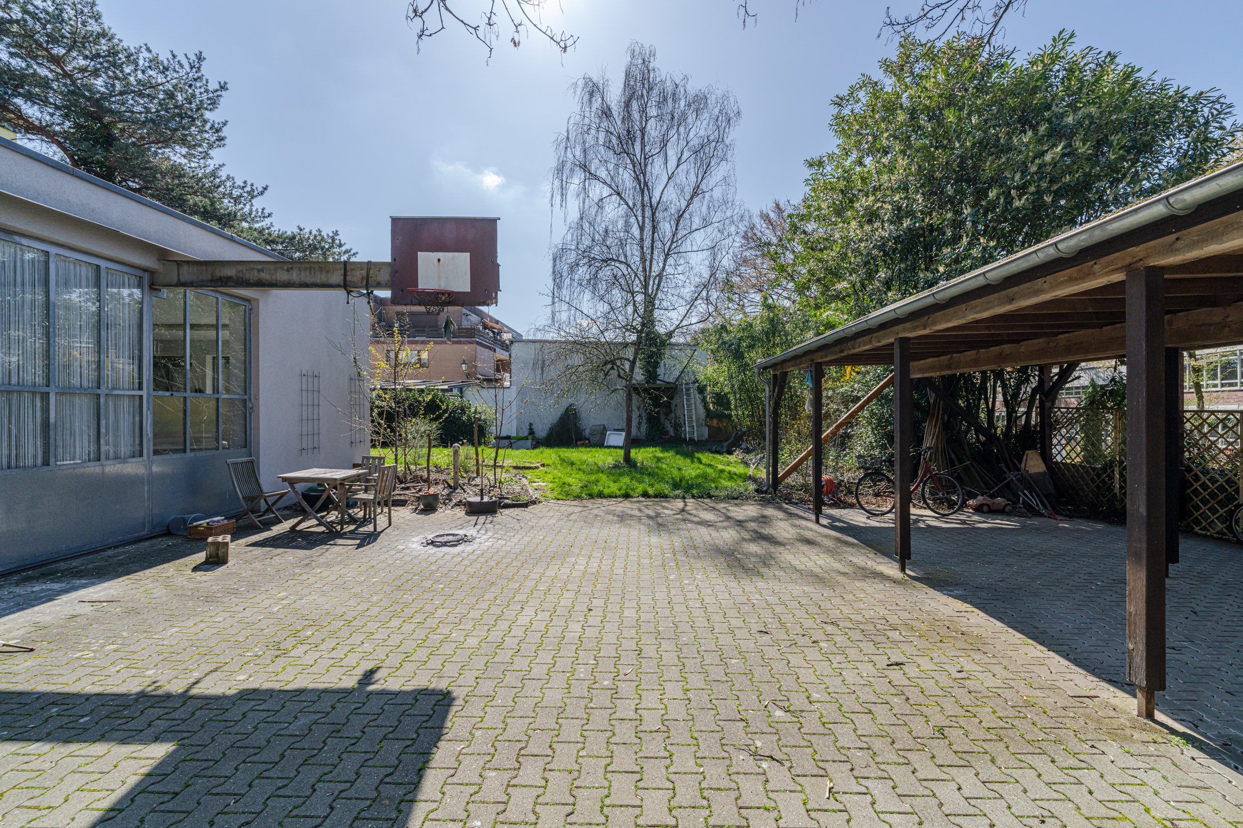 Gepflasterter Hof mit Carport, Basketballkorb, Sitzgruppe und Gartenbereich mit Bäumen.