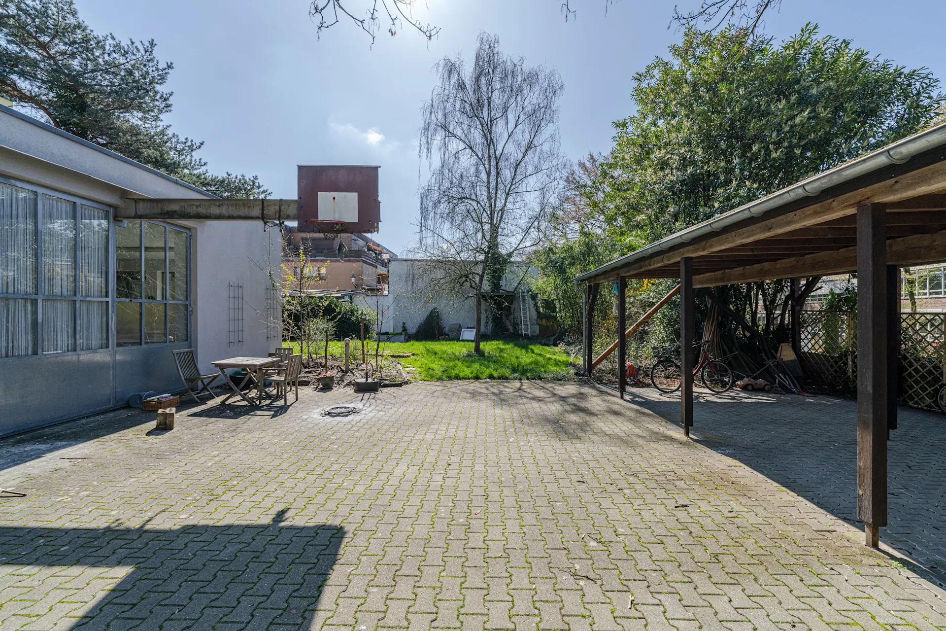 Gepflasterter Hof mit Carport, Basketballkorb, Sitzgruppe und Gartenbereich mit Bäumen.