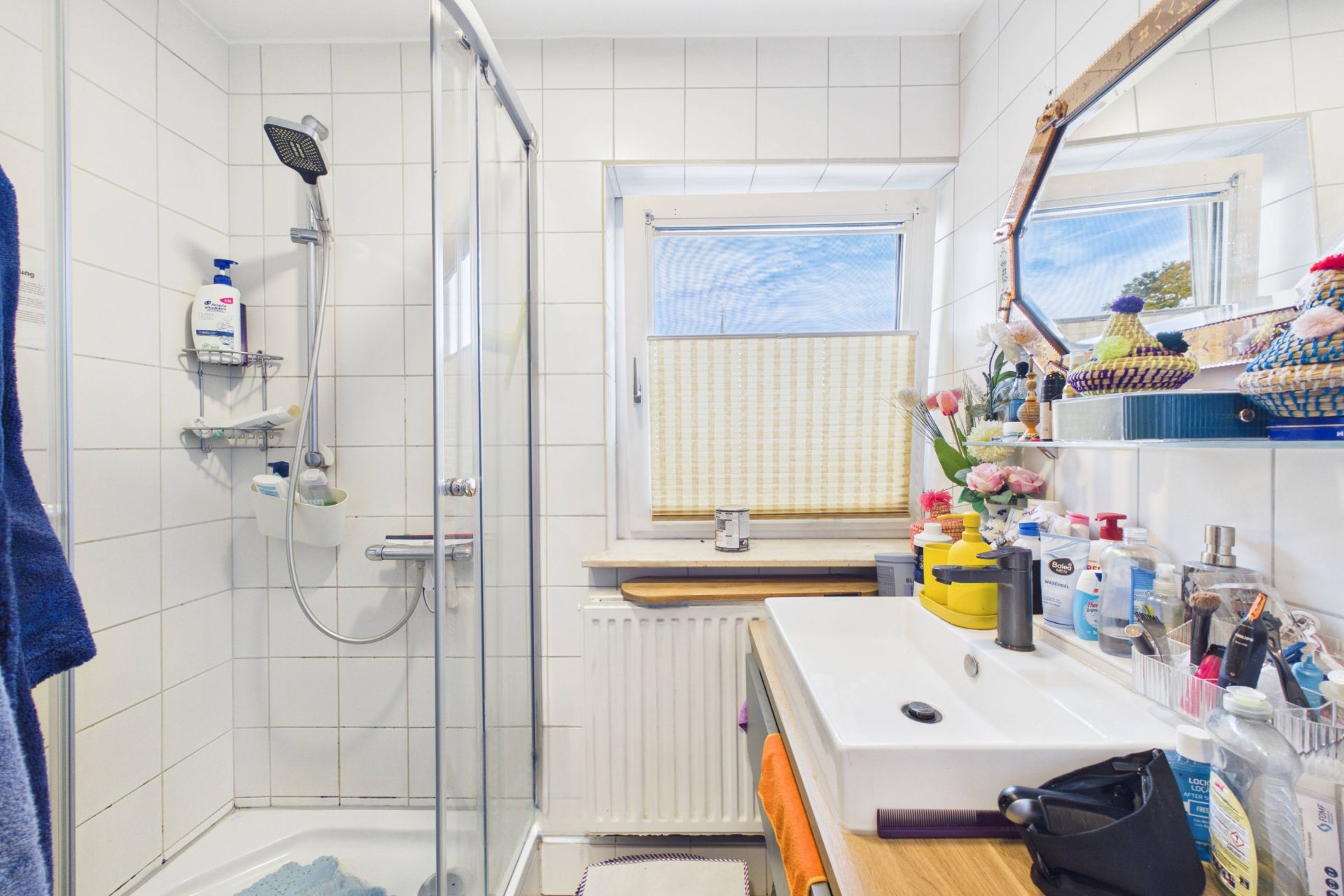 Kleines Badezimmer mit Dusche, Waschbecken, Spiegel und Fenster, viele Pflegeartikel auf dem Waschbecken.