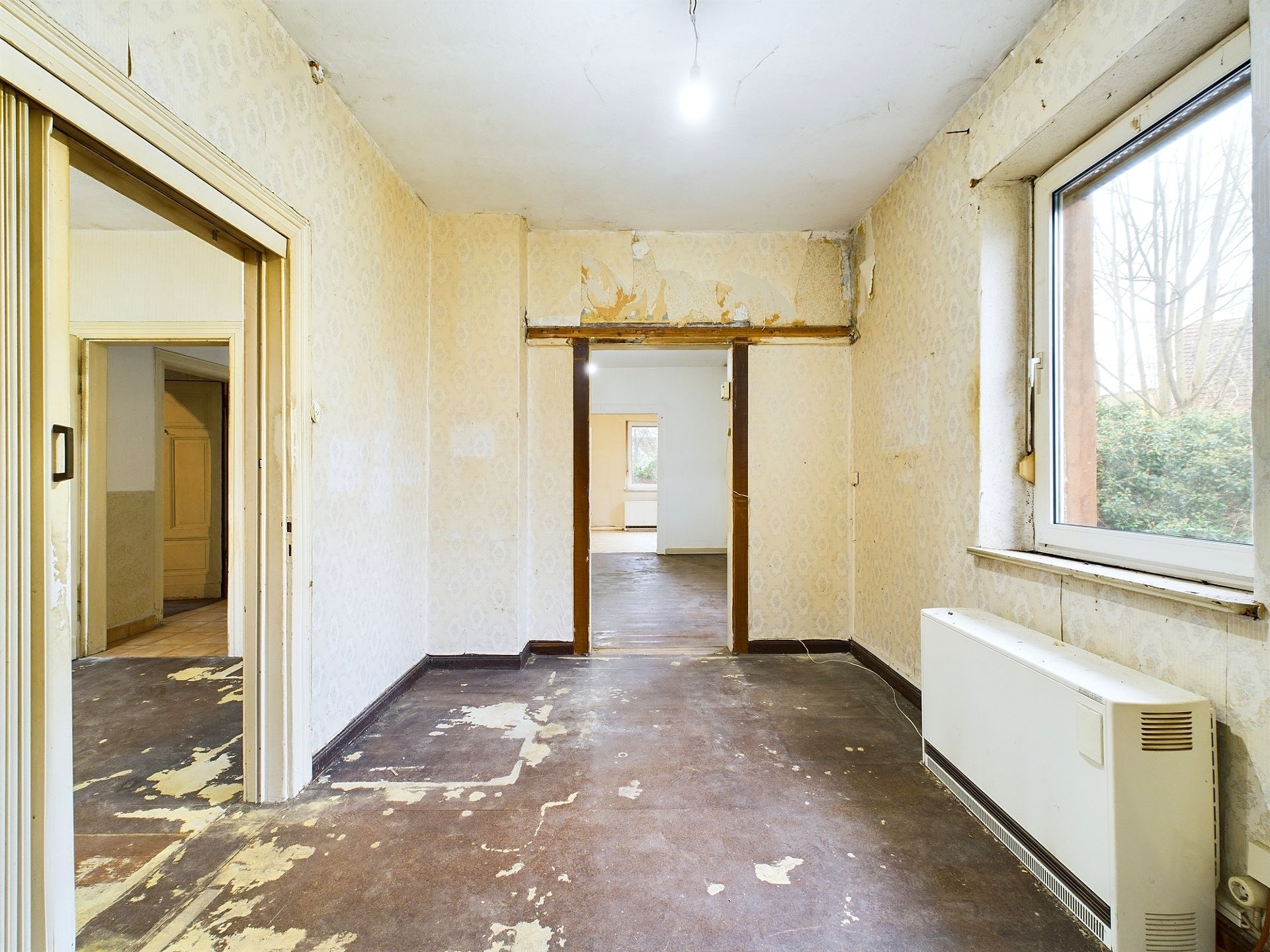 Altbau-Zimmer mit abgenutztem Boden, Tapeten und großem Fenster, Blick in weitere Räume.