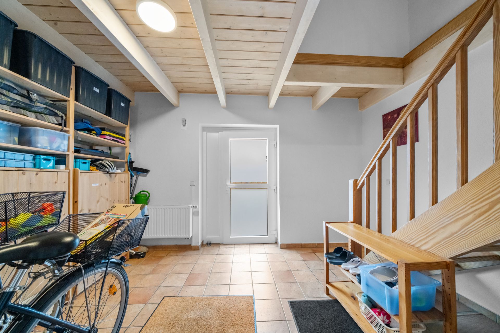 Flur mit gefliestem Boden, Holztreppe, Regalen und einem Fahrrad, helle Decke mit Holzbalken.