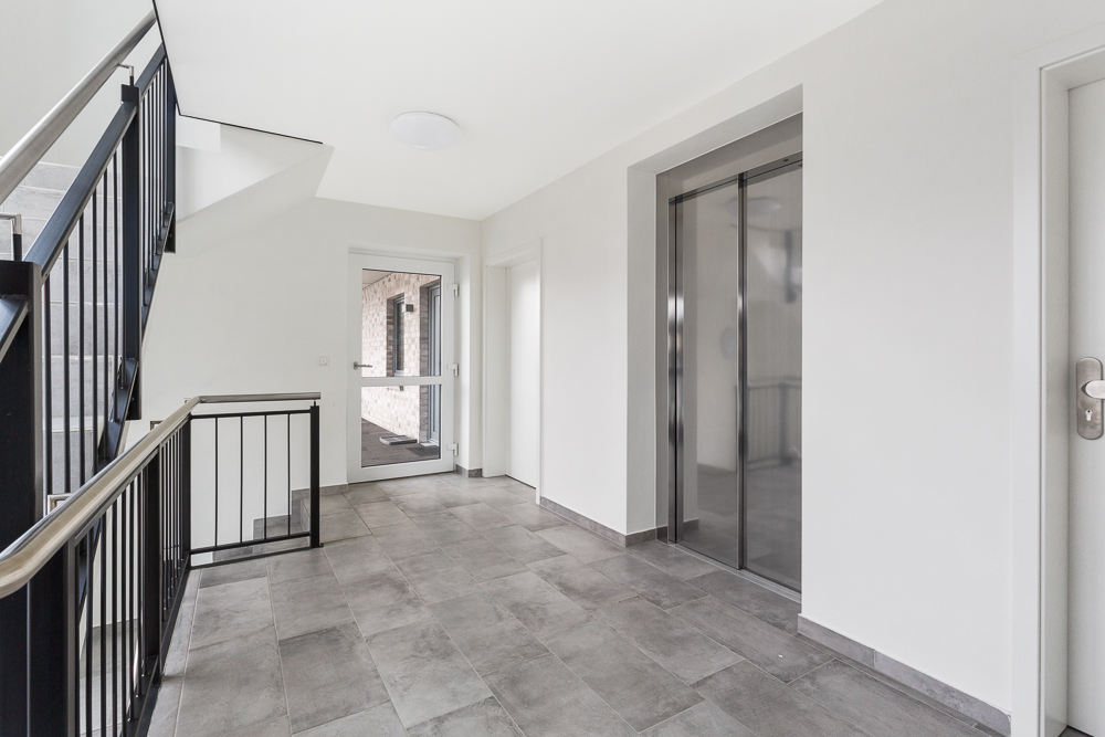 Heller Hausflur mit Aufzug, Treppe und Glastür, graue Bodenfliesen.