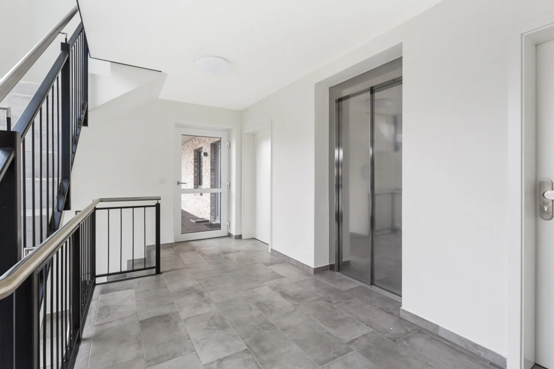 Heller Hausflur mit Aufzug, Treppe und Glastür, graue Bodenfliesen.