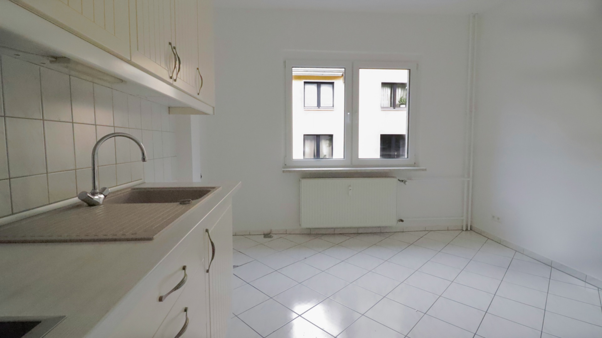 Helle, leere Küche mit Fliesenboden, Spüle, weißen Schränken und Fenster.