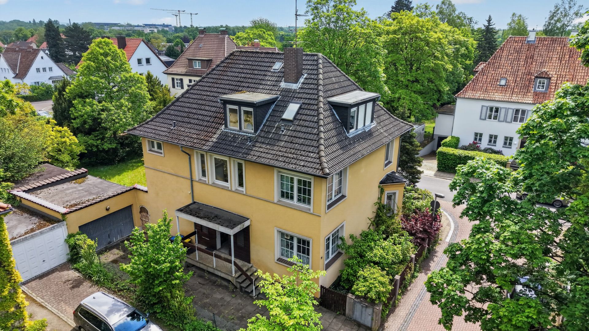 Gelbes, freistehendes Wohnhaus mit dunklem Dach, umgeben von Bäumen und Nachbarhäusern.