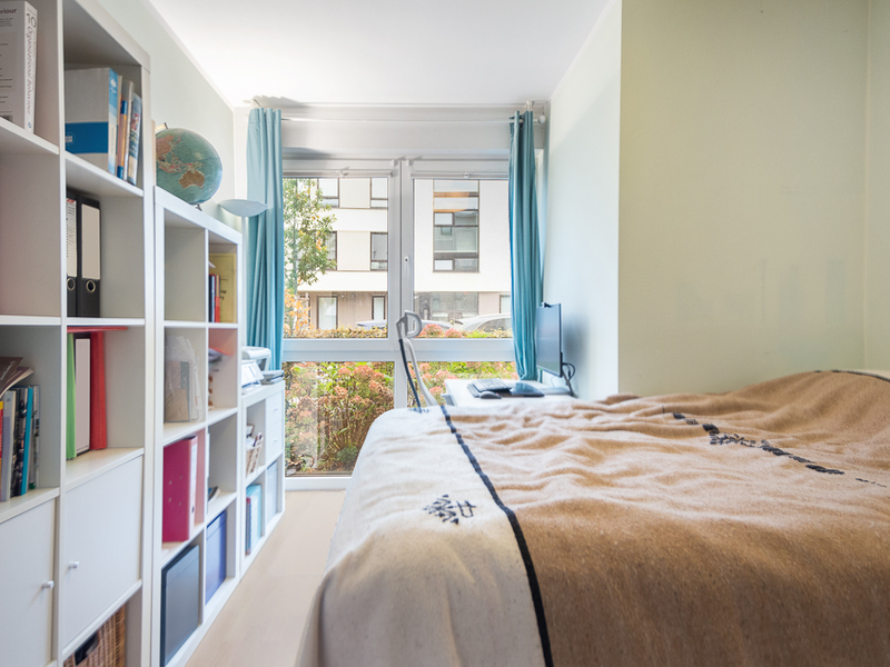 Helles Schlafzimmer mit Bett, Regal, Schreibtisch und großem Fenster mit blauen Vorhängen.