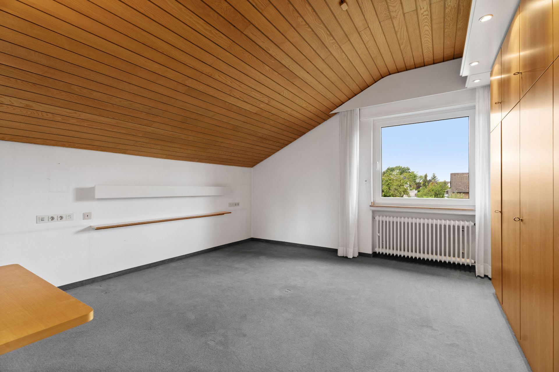 Leeres Zimmer mit Holzdecke, grauem Teppich, großem Fenster und Einbauschränken.