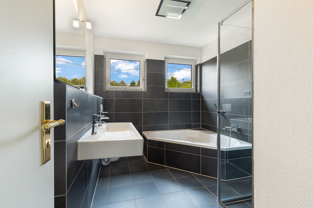 Modernes Badezimmer mit Eckbadewanne, Dusche, Waschbecken und drei Fenstern.