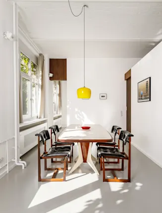 Heller Essbereich mit Holztisch, sechs Stühlen, gelber Lampe und großen Fenstern.