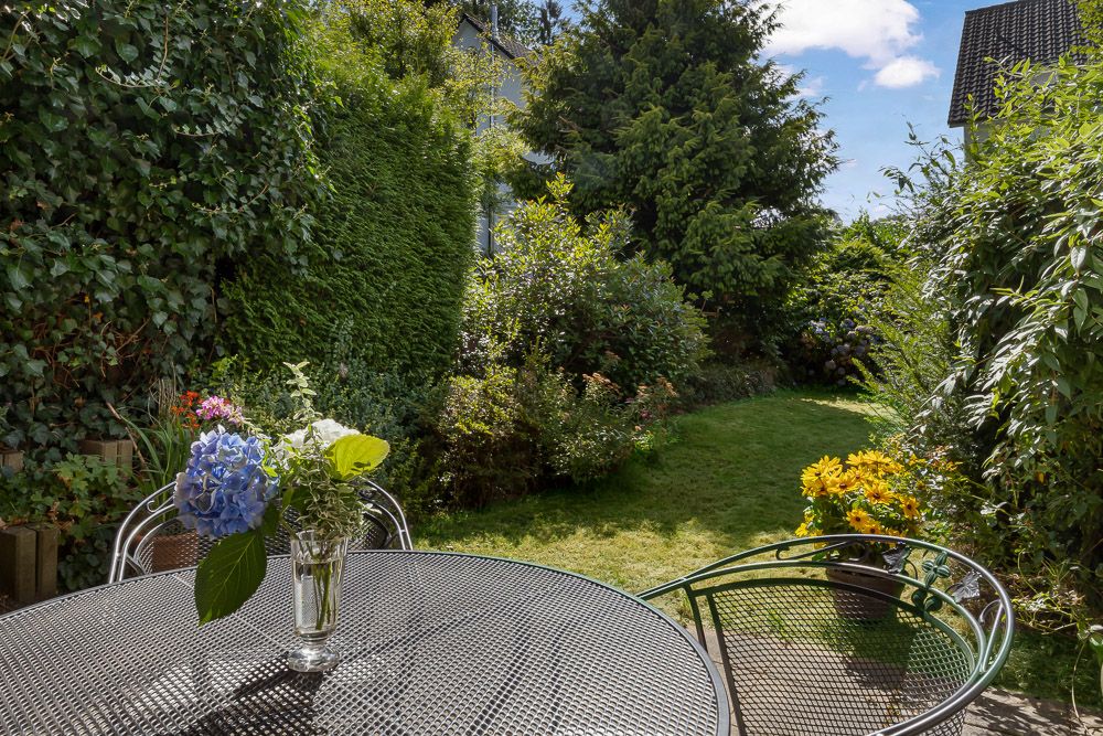 Garten mit rundem Metalltisch, Blumenstrauß und Stühlen, umgeben von grünen Büschen und Rasen.