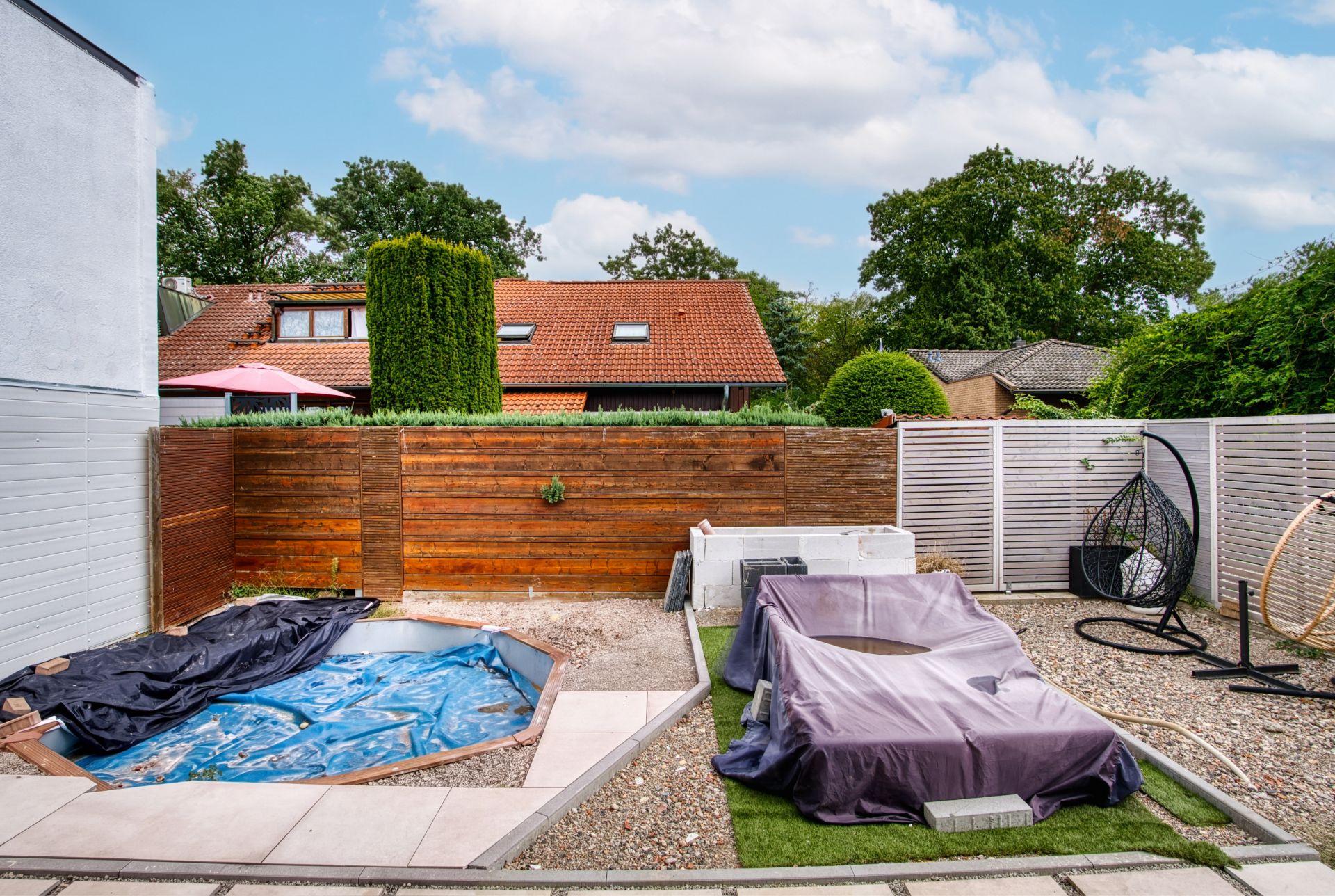 Gartenbereich mit abgedecktem Pool und abgedeckten Gartenmöbeln, Holzzaun im Hintergrund.