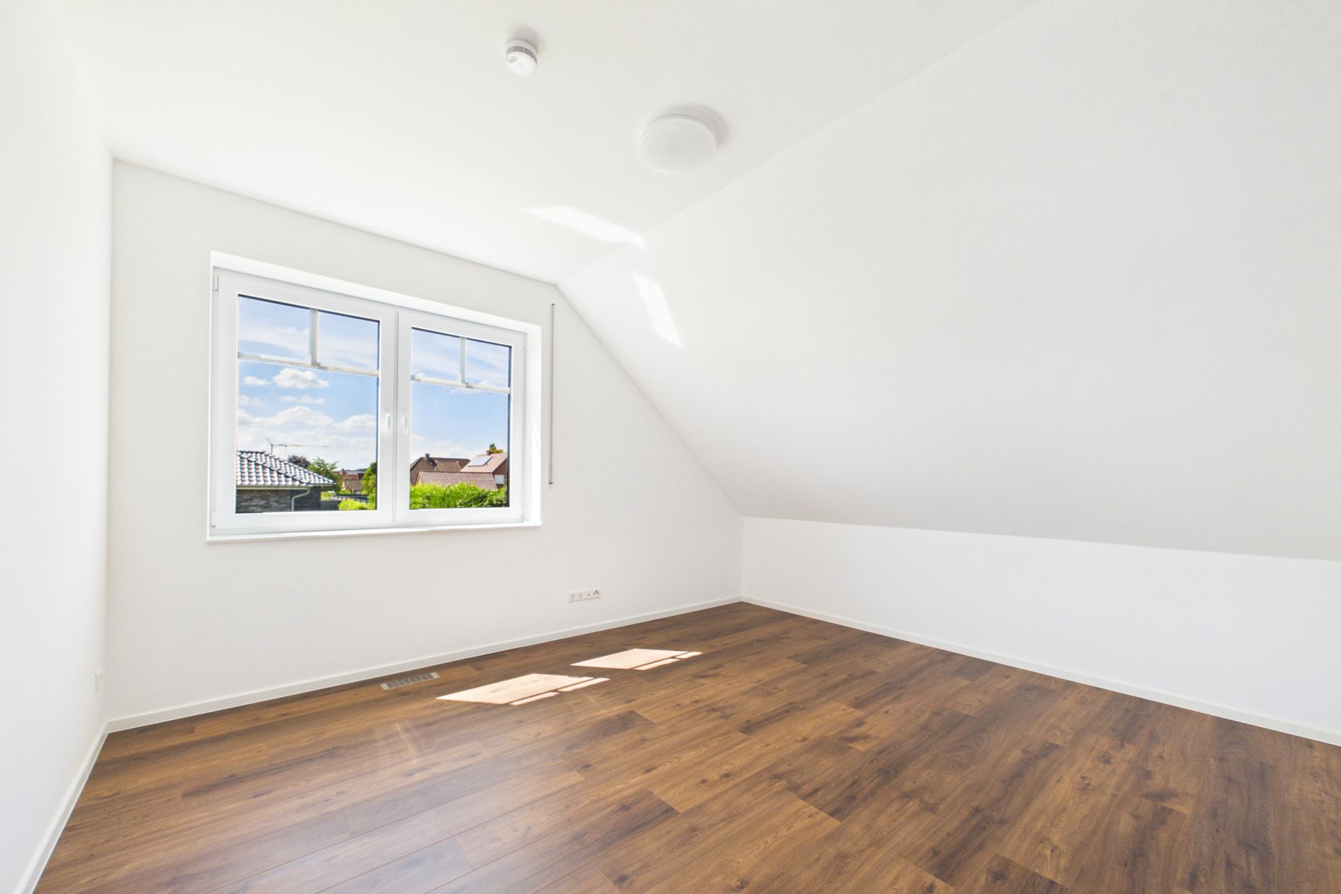 Helles, leeres Zimmer mit schräger Decke, Holzfußboden und Fenster mit Ausblick.