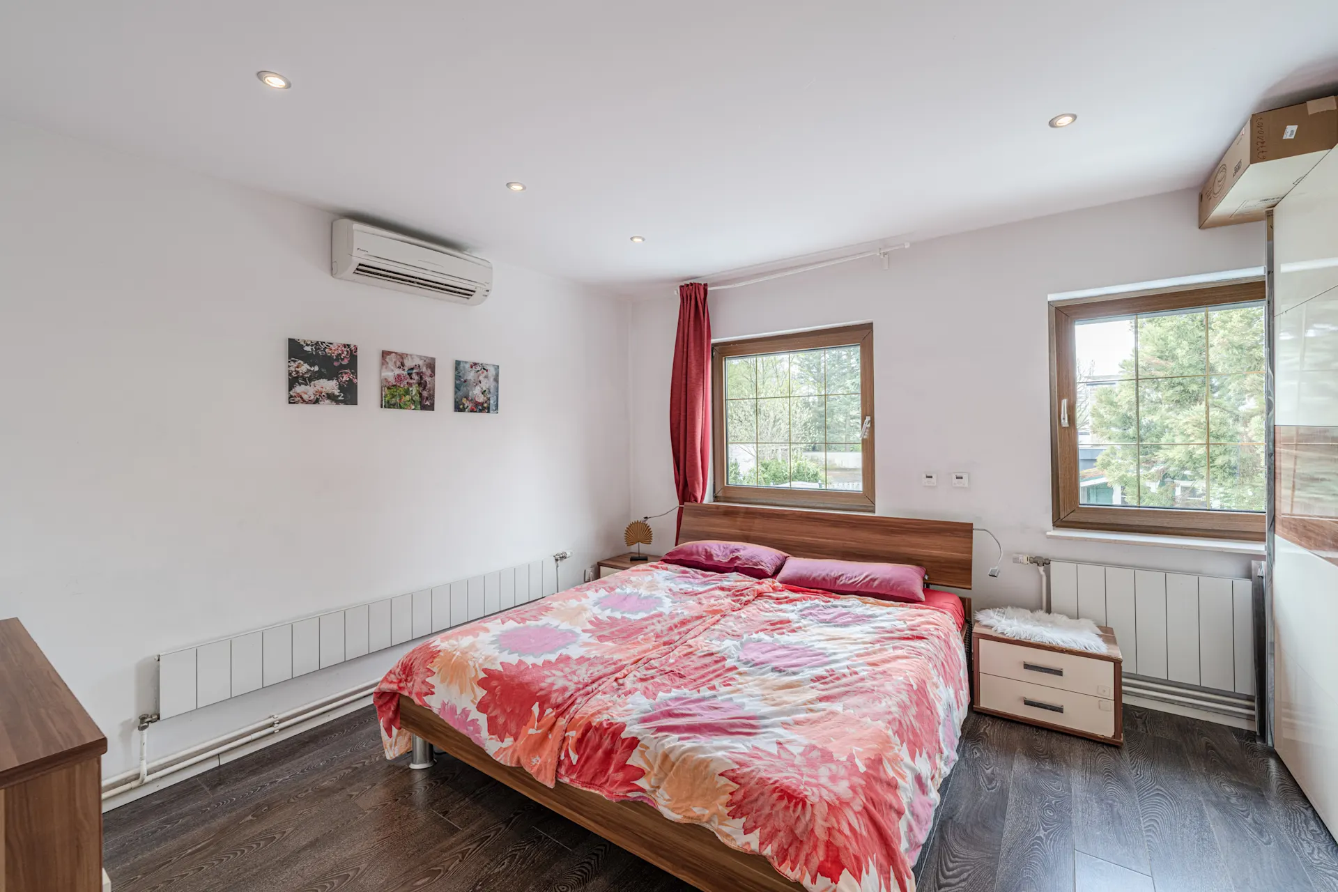 Schlafzimmer mit Doppelbett, buntem Bettbezug, zwei Fenstern und Wandklimaanlage.