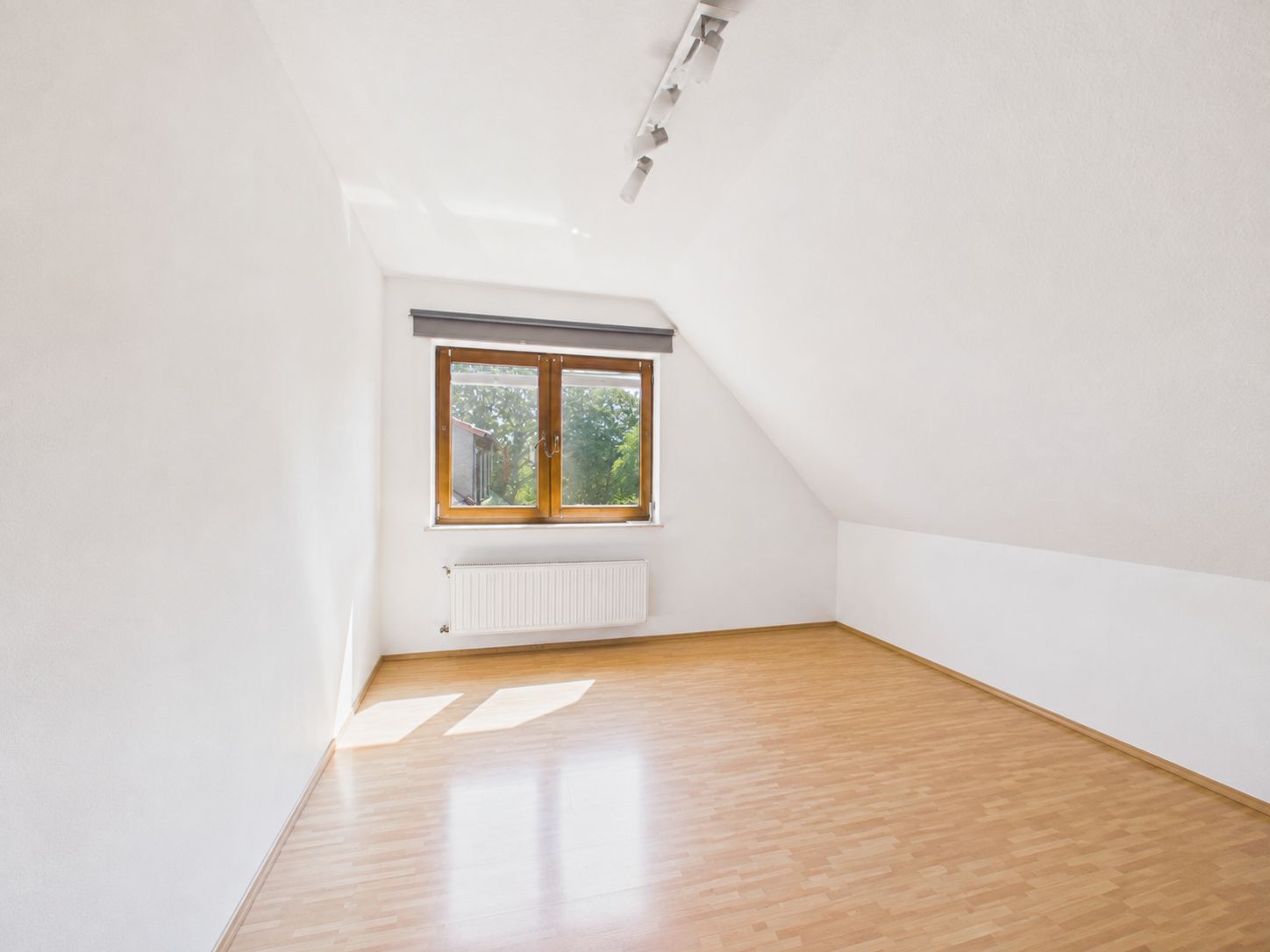 Helles, leeres Zimmer mit schräger Decke, Holzboden und Fenster mit Blick ins Grüne.