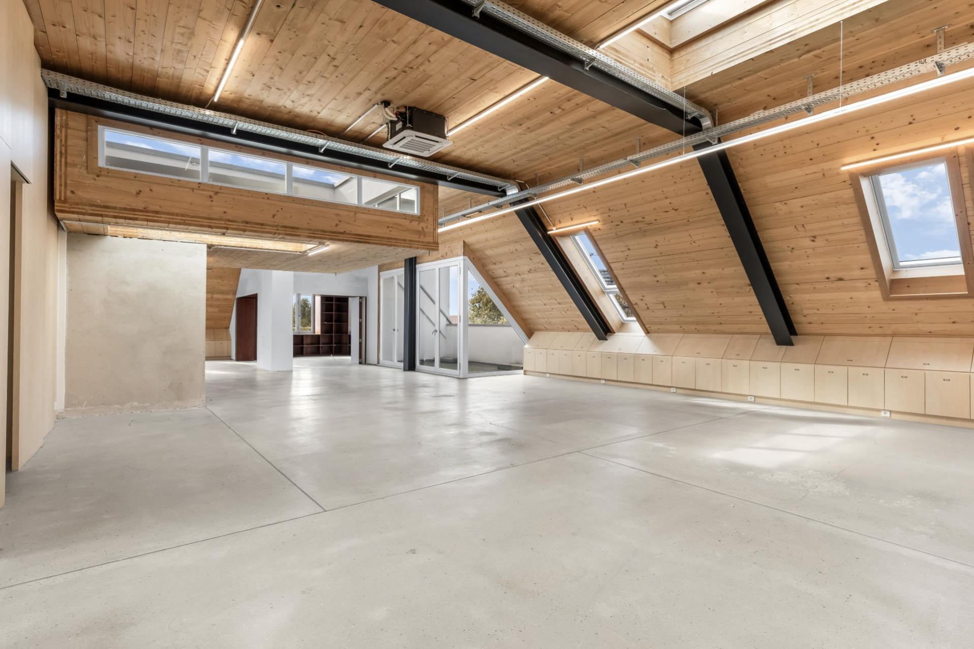 Heller Raum mit schrägen Holzdecken, großen Fenstern und sichtbaren schwarzen Trägern.
