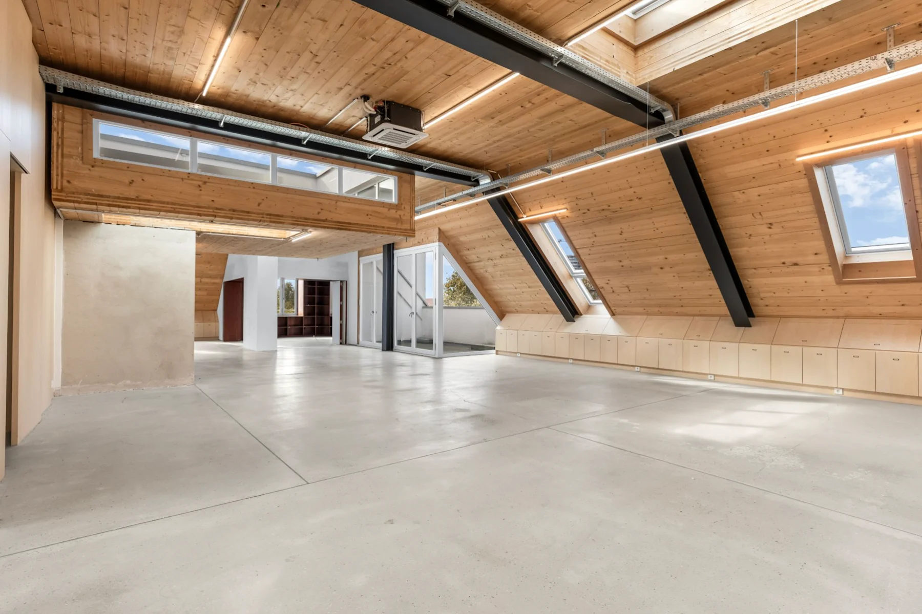 Heller Raum mit schrägen Holzdecken, großen Fenstern und sichtbaren schwarzen Trägern.