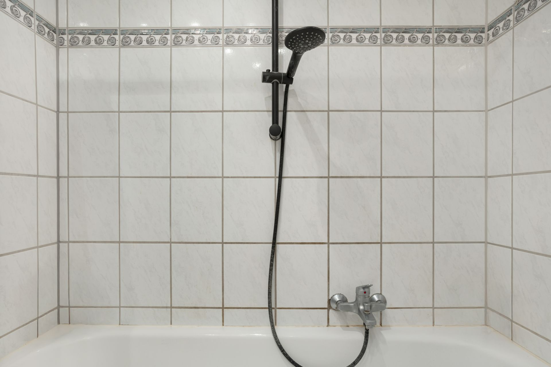 Weiße, geflieste Badewanne mit Duschkopf und Armatur vor gemusterter Fliesenwand.