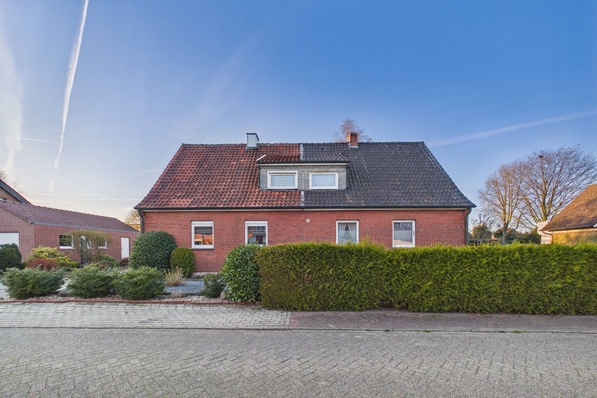Backsteinhaus mit rotem Dach, Hecken und Vorgarten an einer ruhigen Straße.