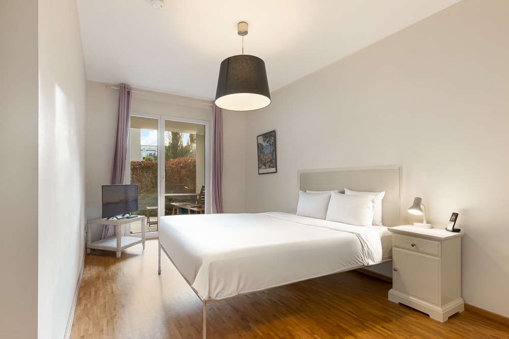 Helles Schlafzimmer mit Doppelbett, Nachttisch, Fernseher und Zugang zu einer Terrasse.