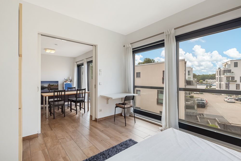 Helles Zimmer mit großen Fenstern, kleinem Schreibtisch und Blick auf moderne Wohngebäude.