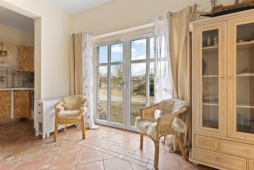 Heller Raum mit zwei Korbstühlen, Glasschrank und großem Fenster mit Blick nach draußen.