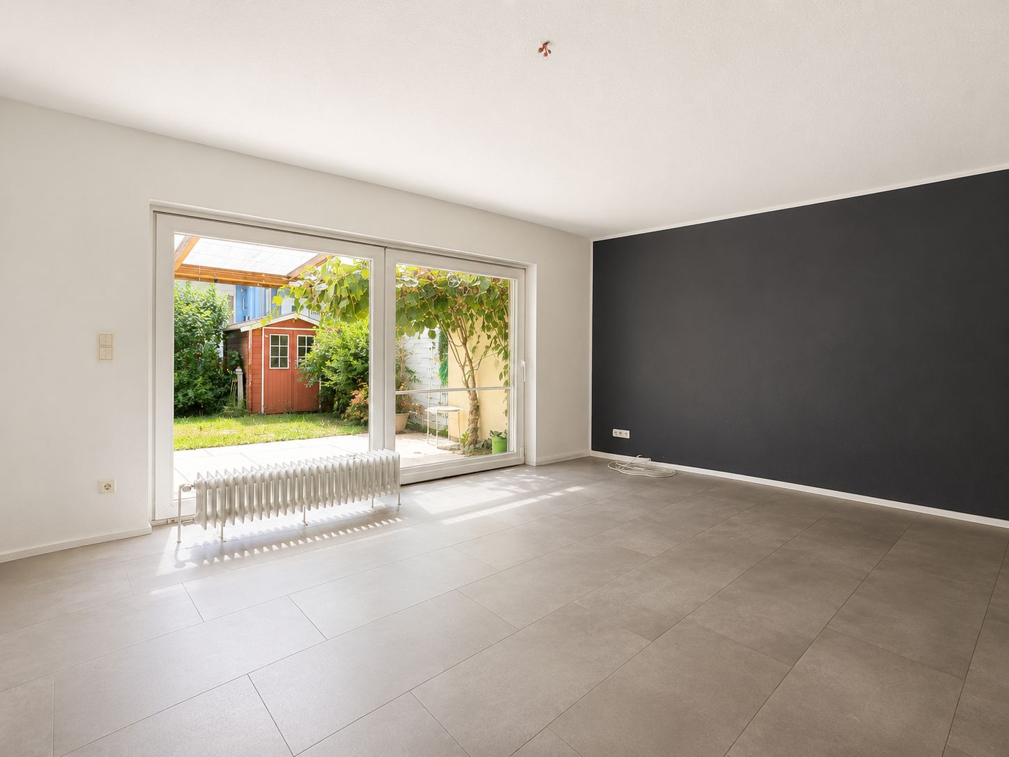 Leeres Wohnzimmer mit grauer Wand, großen Fenstern und Blick in den Garten mit Gartenhaus.