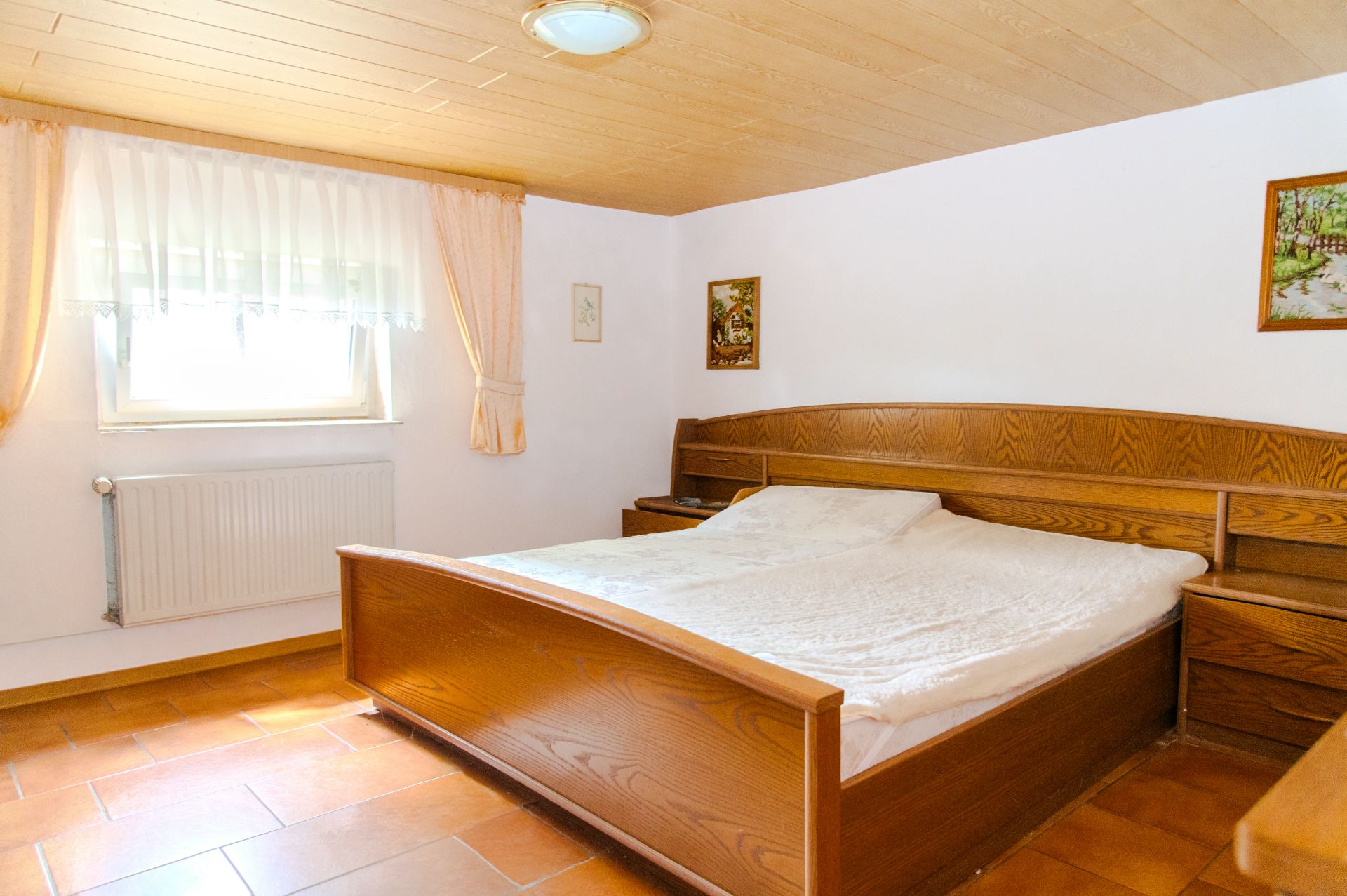 Schlafzimmer mit großem Holzbett, Nachttischen, Fenster mit Vorhängen und Wandbildern.