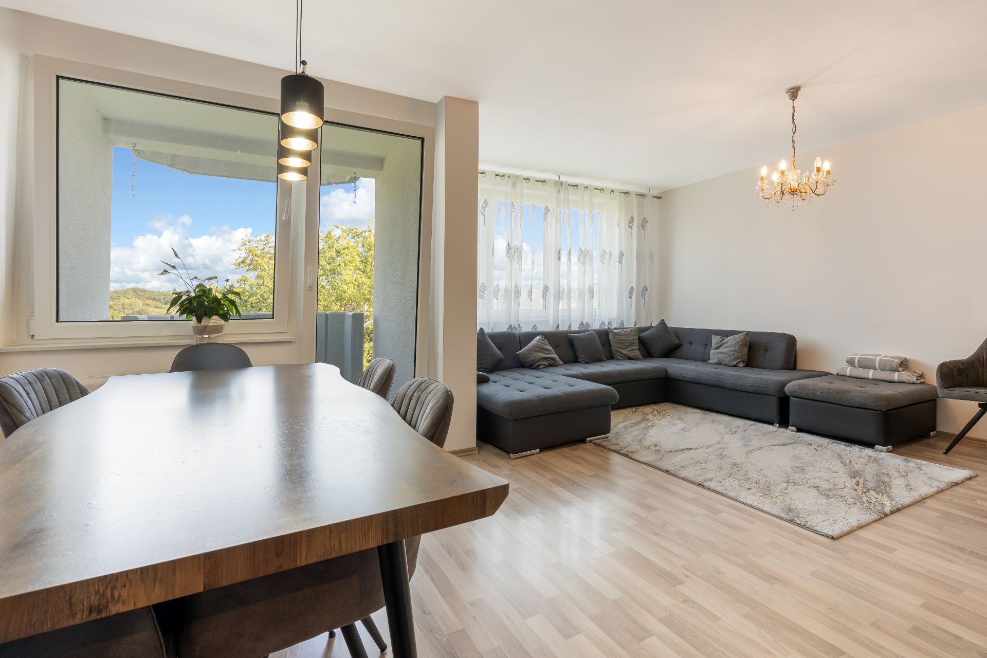 Helles Wohnzimmer mit grauem Ecksofa, Esstisch, großem Fenster und Kronleuchter.