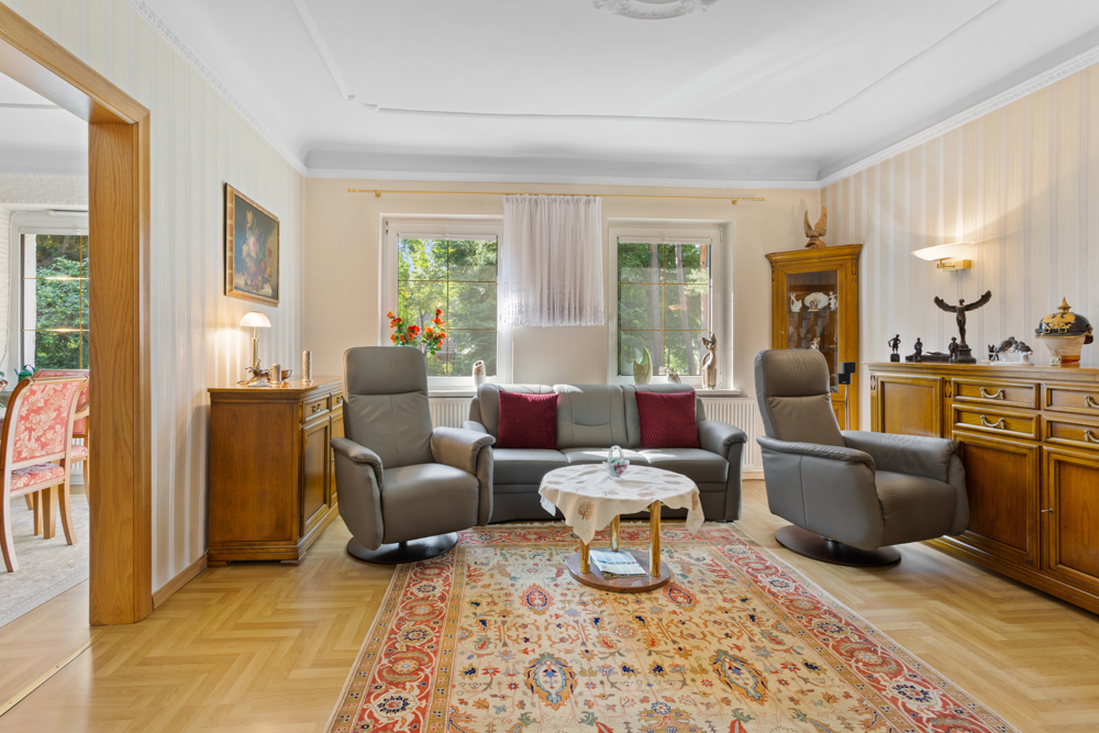 Wohnzimmer mit Sofa, zwei Sesseln, Teppich und Holzmöbeln, zwei Fenster mit Blick ins Grüne.
