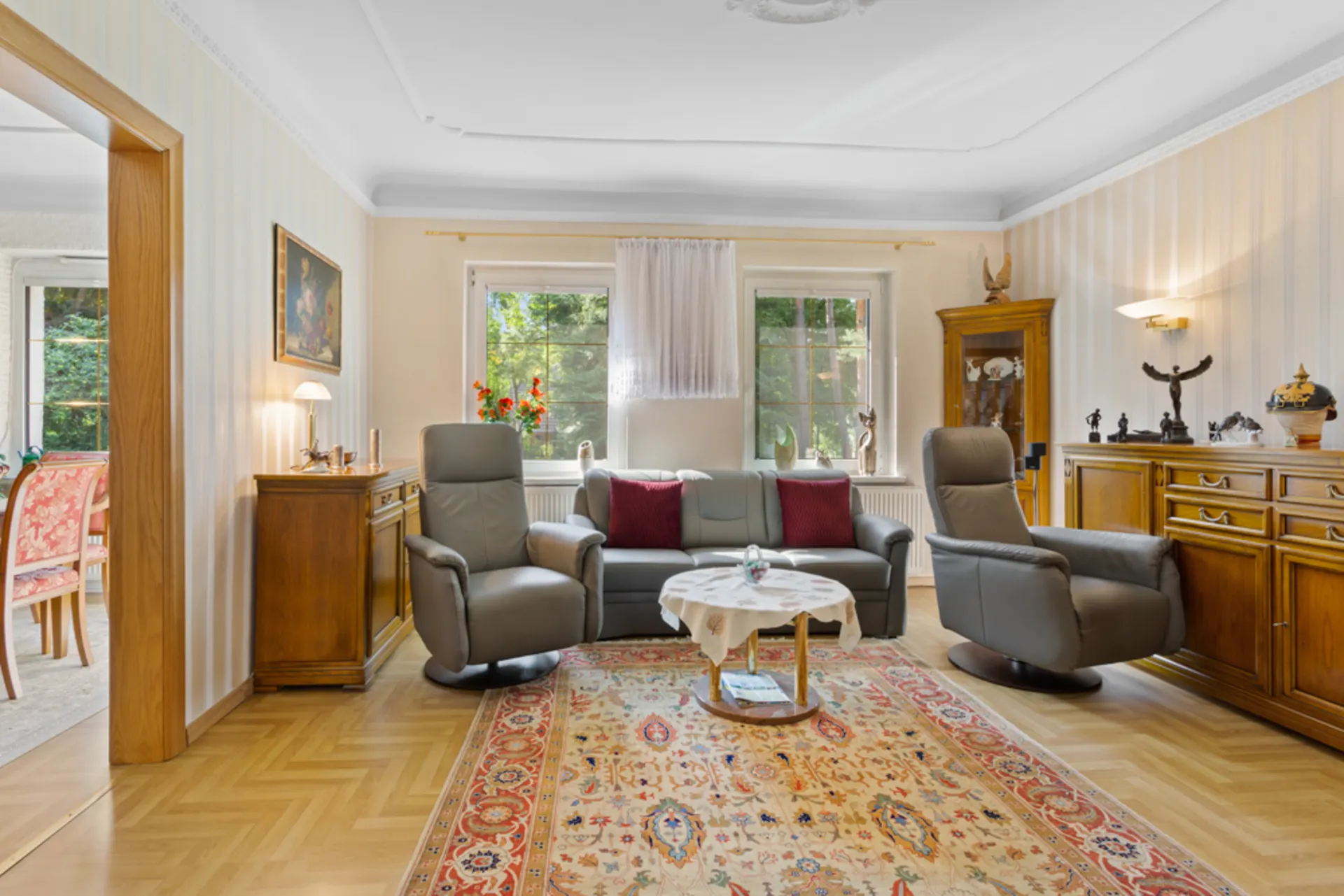Wohnzimmer mit Sofa, zwei Sesseln, Teppich und Holzmöbeln, zwei Fenster mit Blick ins Grüne.