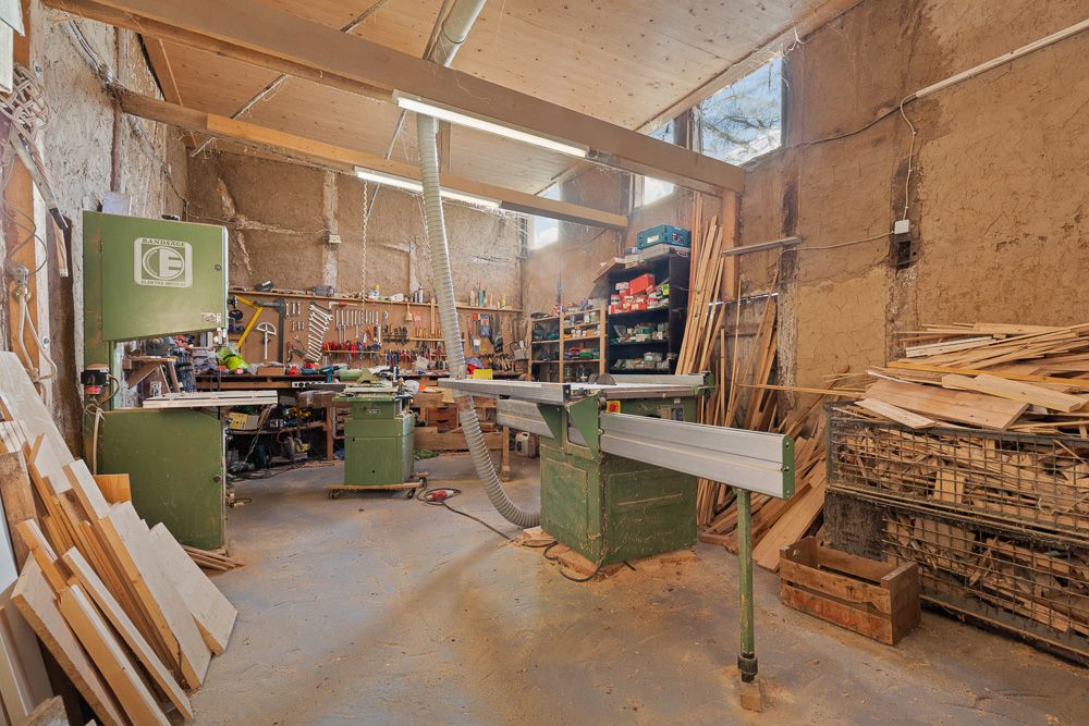 Helle Werkstatt mit Holzbalken, Sägemaschine, Werkbänken, Holzstapel und Regalen voller Werkzeuge.