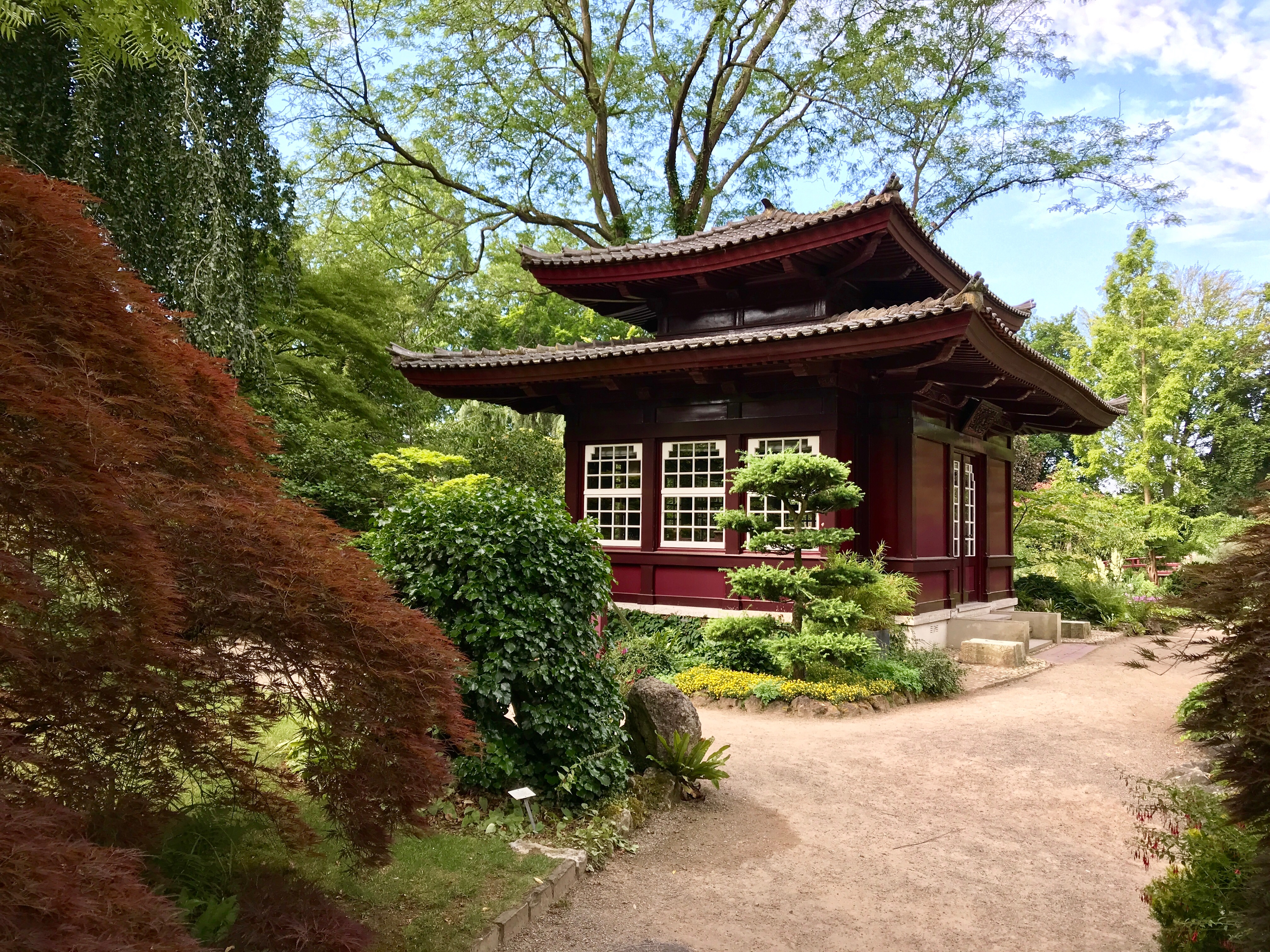 Rotes asiatisches Gartenhaus mit Pavillondach, umgeben von Gartenpflanzen und Bäumen.