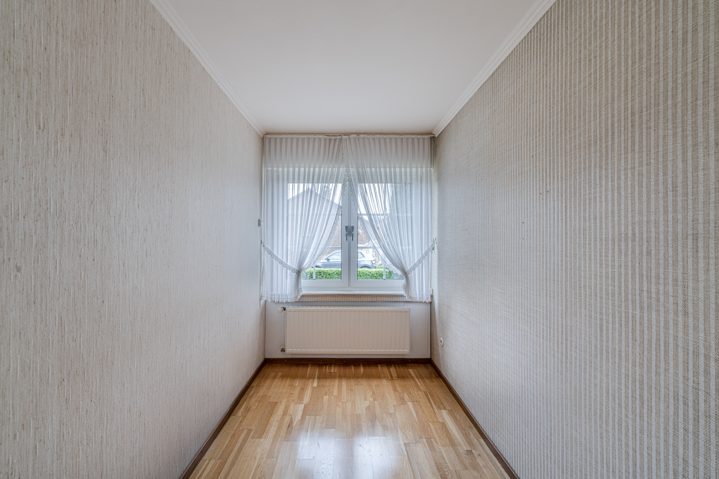 Schmaler Raum mit Holzboden, Fenster mit Gardinen und darunter einem Heizkörper.