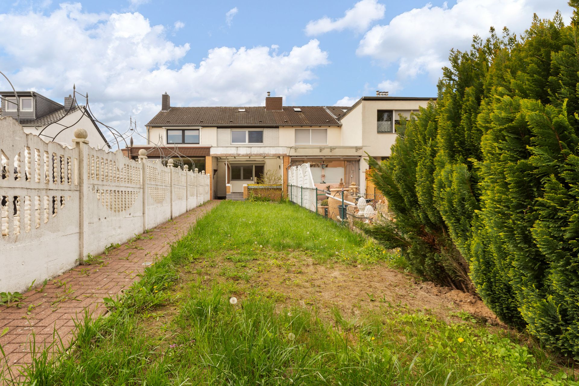 Garten mit Rasen, Weg, Zaun und Reihenhaus im Hintergrund bei Tageslicht.