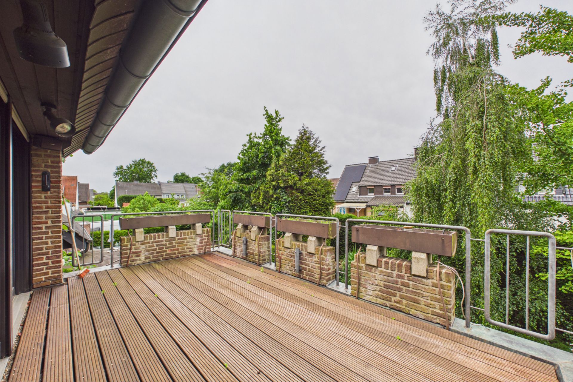 Große Holzterrasse mit Geländer, Blick auf Bäume und Nachbarhäuser.