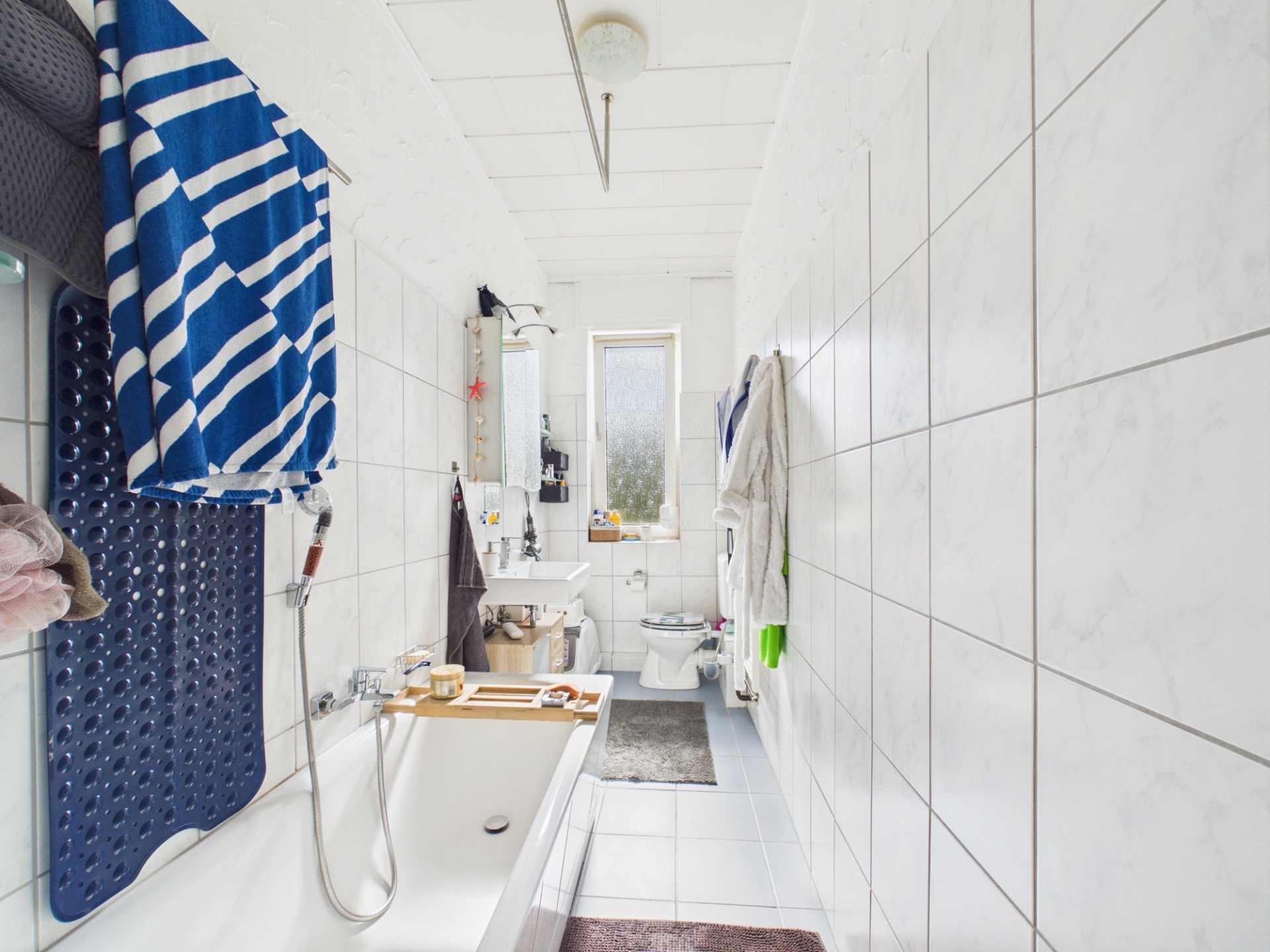 Längliches Badezimmer mit Badewanne, Waschbecken, WC und Fenster am Ende des Raumes.