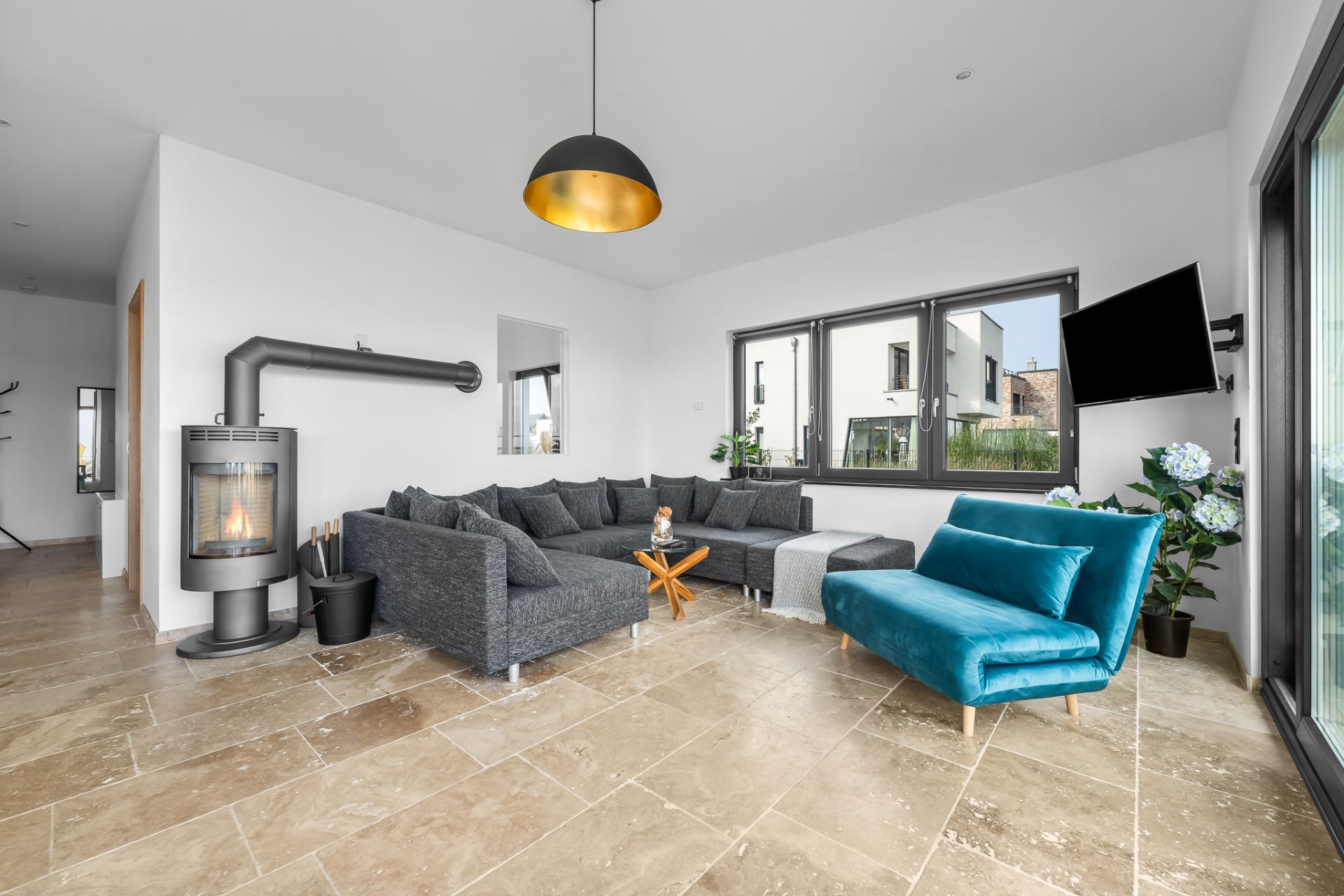 Modernes Wohnzimmer mit Ecksofa, blauem Sessel, Kaminofen und großen Fenstern.