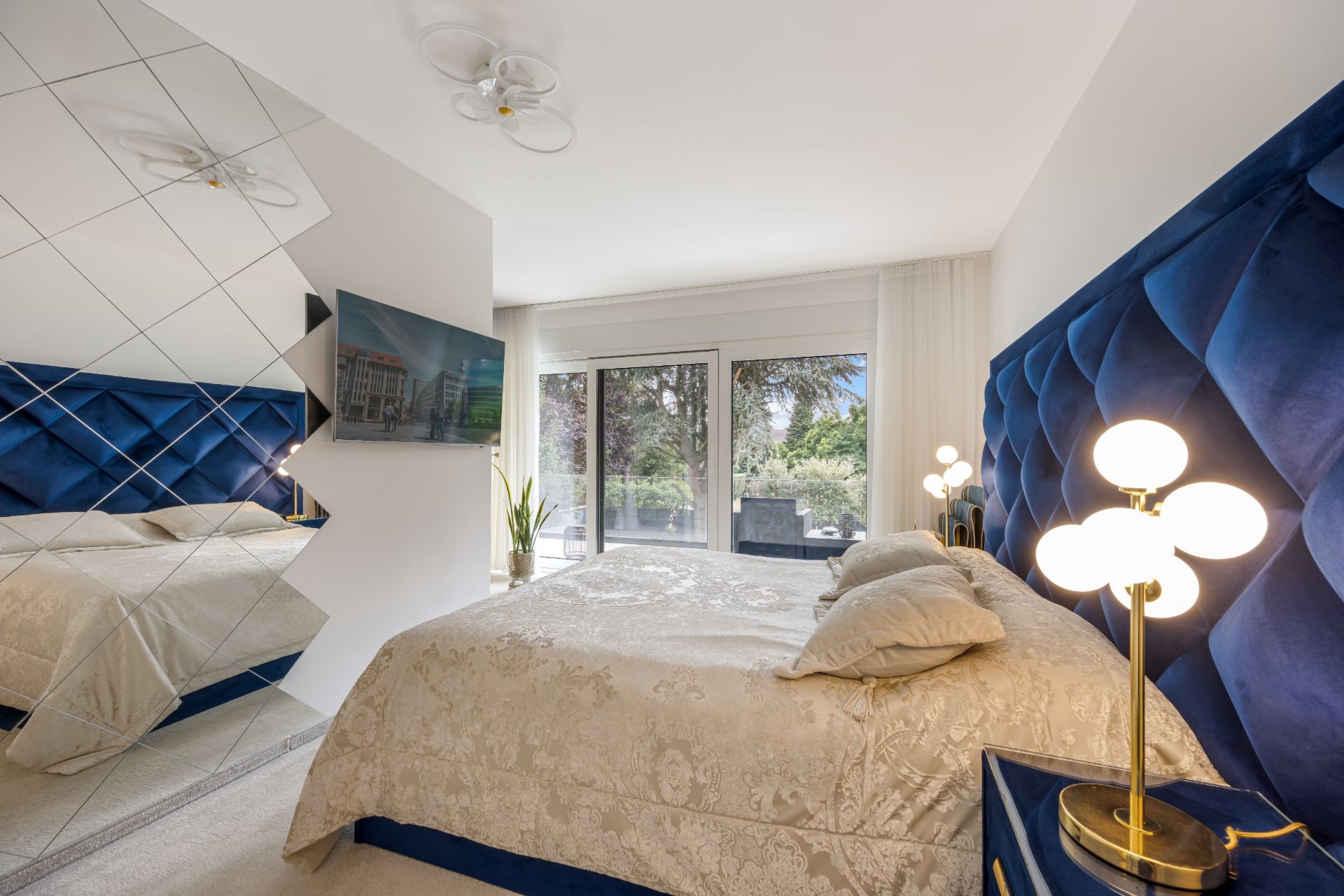 Modernes Schlafzimmer mit blauem Bettkopfteil, Spiegelwand, Fernseher und Zugang zur Terrasse.