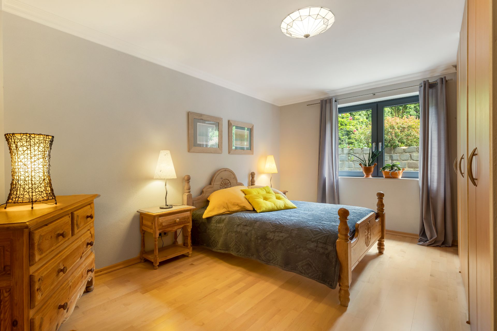 Gemütliches Schlafzimmer mit Holzbett, Kommode, Nachttischlampen und Fenster mit Blick ins Grüne.