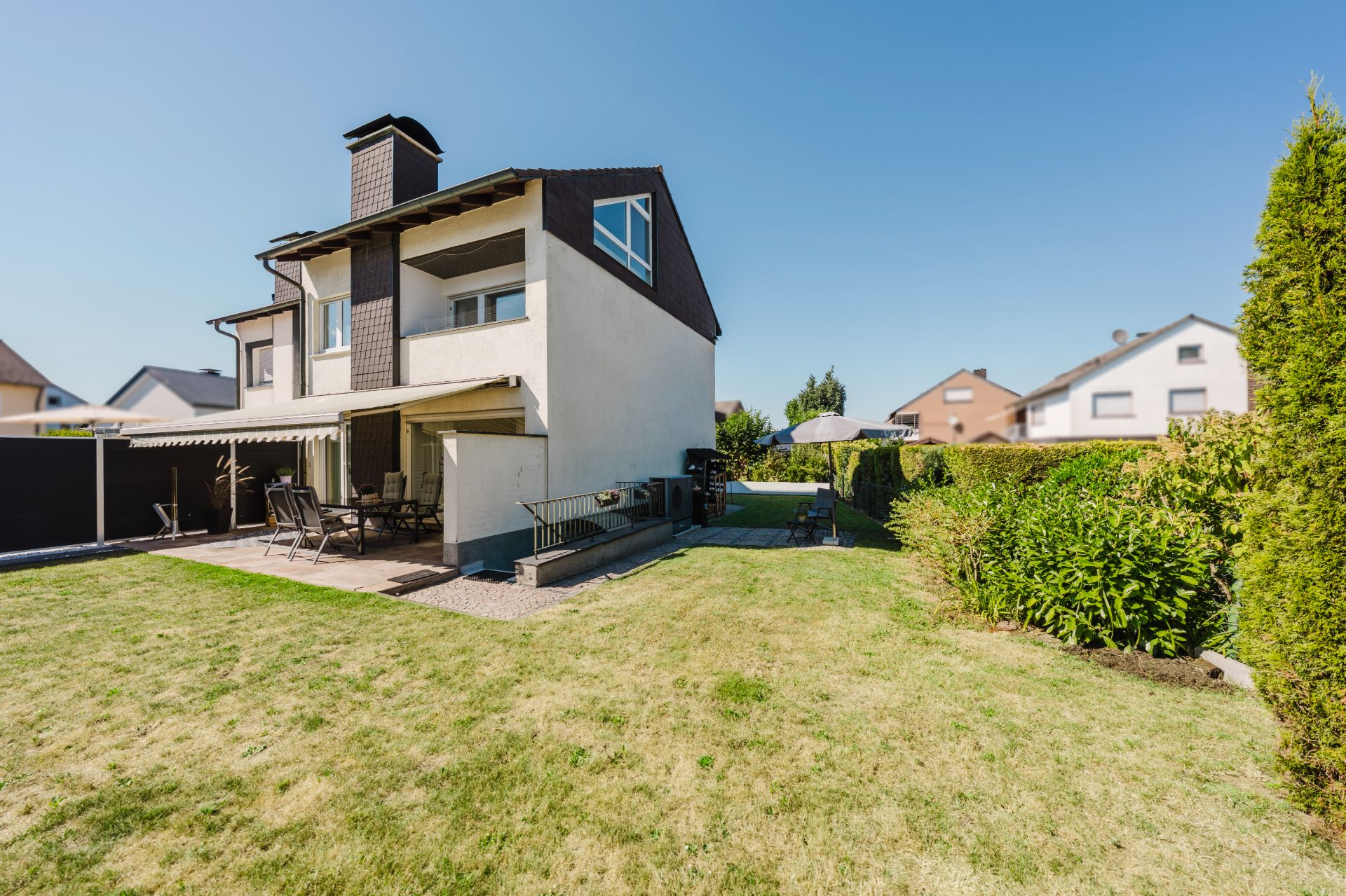 Zweistöckiges Wohnhaus mit Terrasse und Garten, Sonnenschirm und Gartenmöbeln auf Rasenfläche.