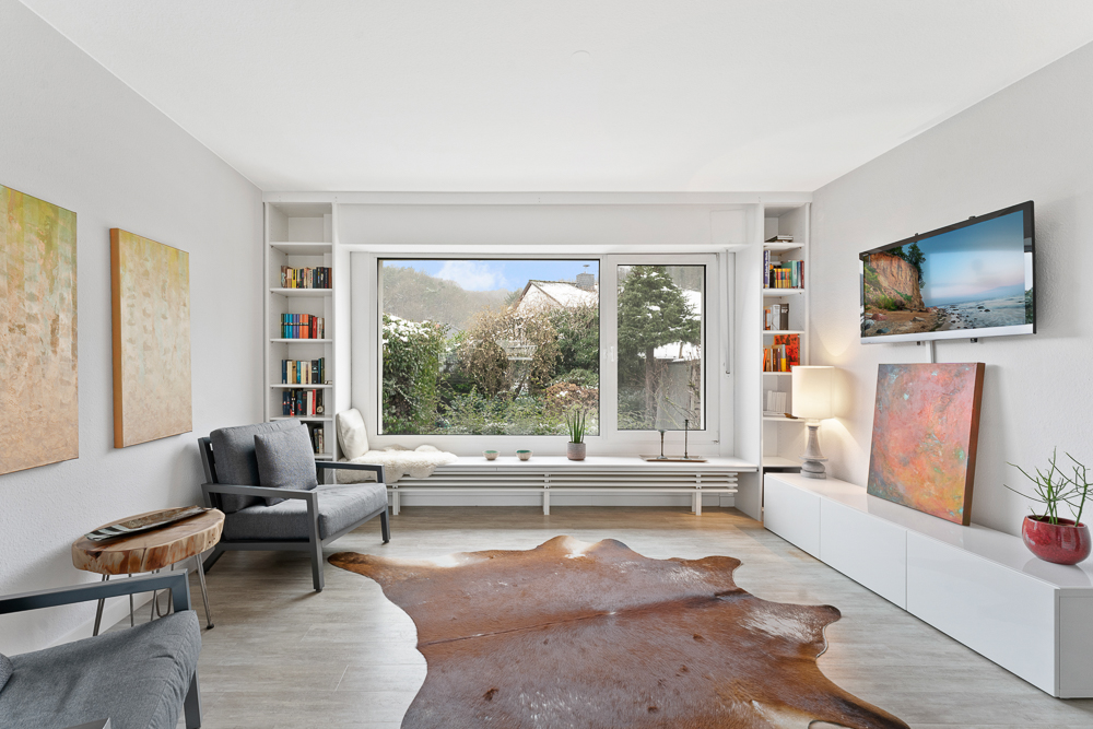 Helles Wohnzimmer mit großem Fenster, Sesseln, Teppich und TV an der Wand.