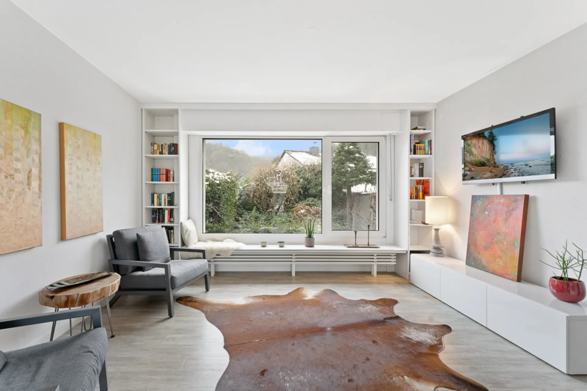 Helles Wohnzimmer mit großem Fenster, Sesseln, Teppich und TV an der Wand.