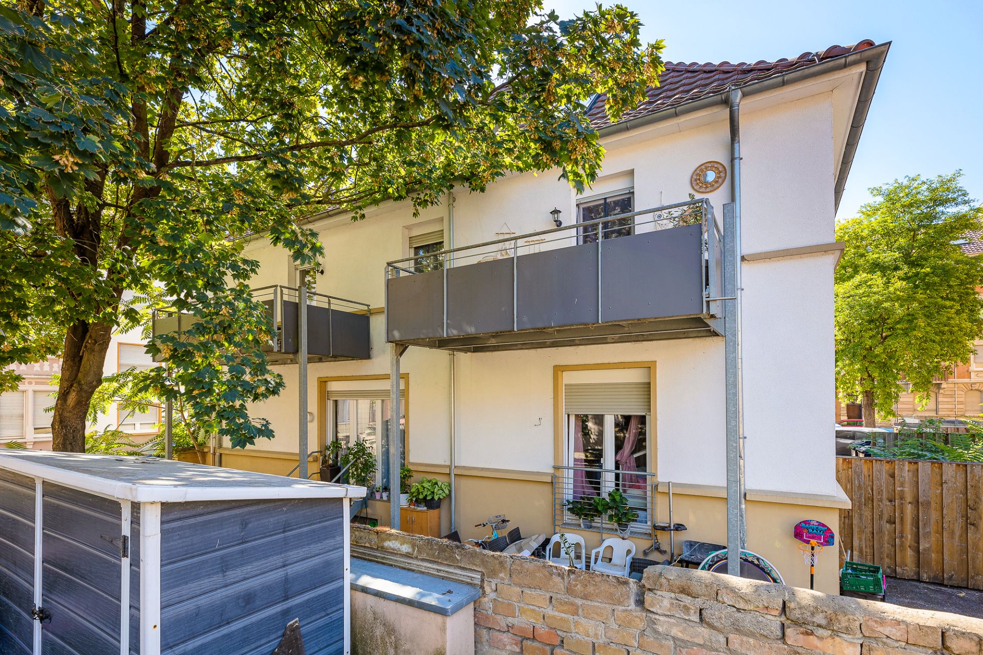 Zweistöckiges Wohnhaus mit Balkon, Sitzbereich im Hof und großem Baum davor.