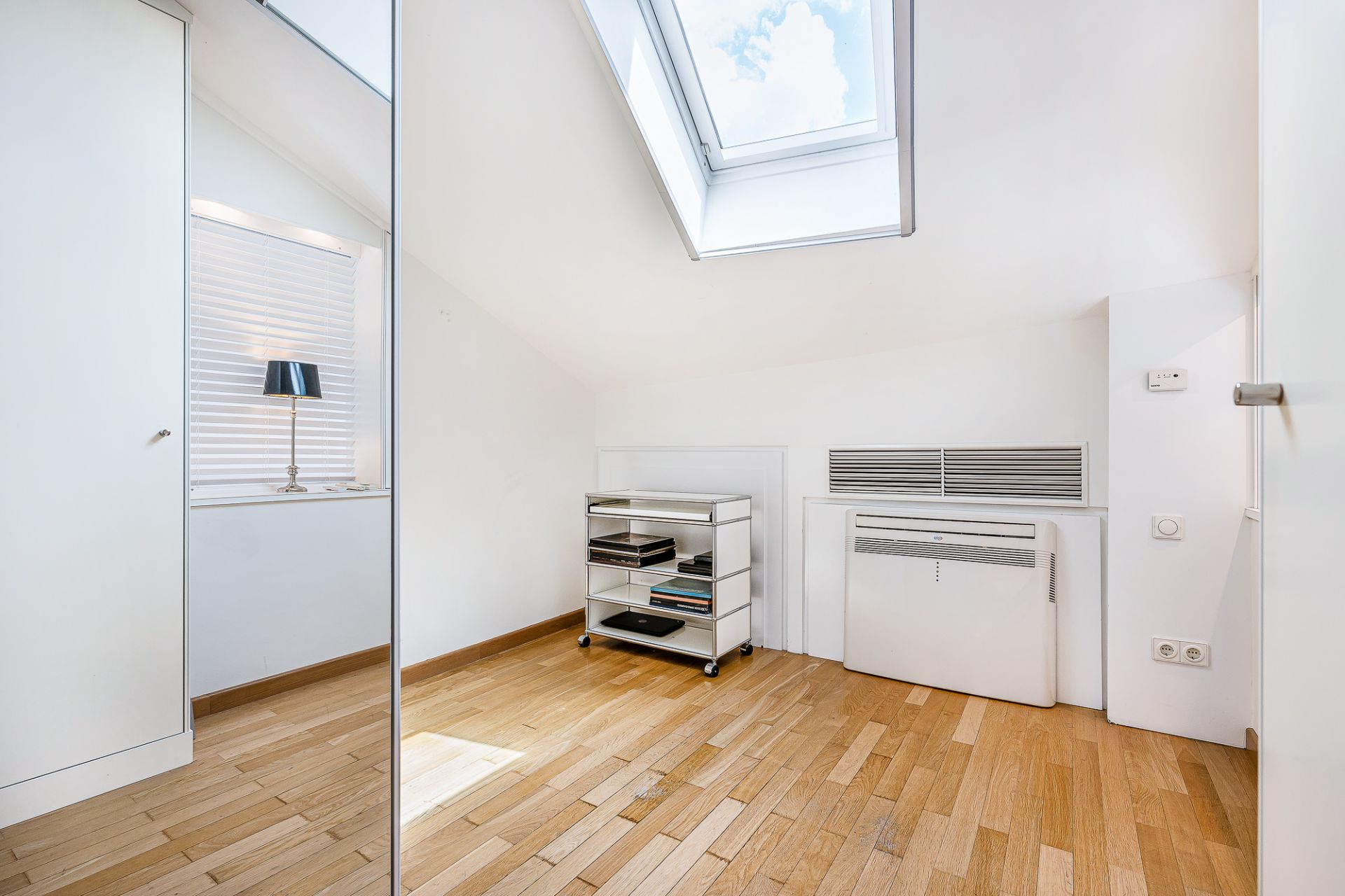 Helles Zimmer mit Holzfußboden, Dachfenster und kleinem Regal auf Rollen.