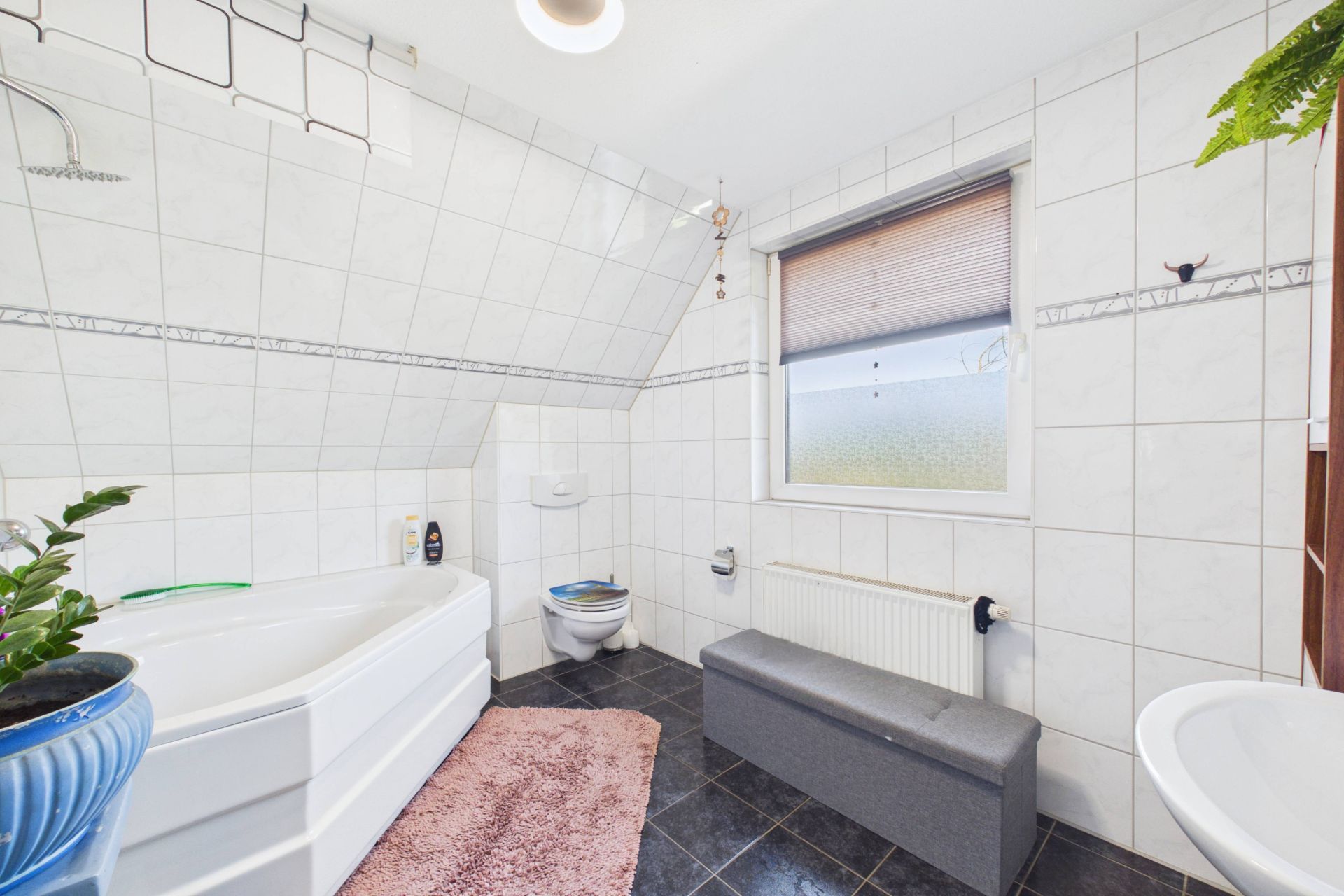 Modernes Badezimmer mit Badewanne, Toilette, Fenster, Bank und Pflanzen.