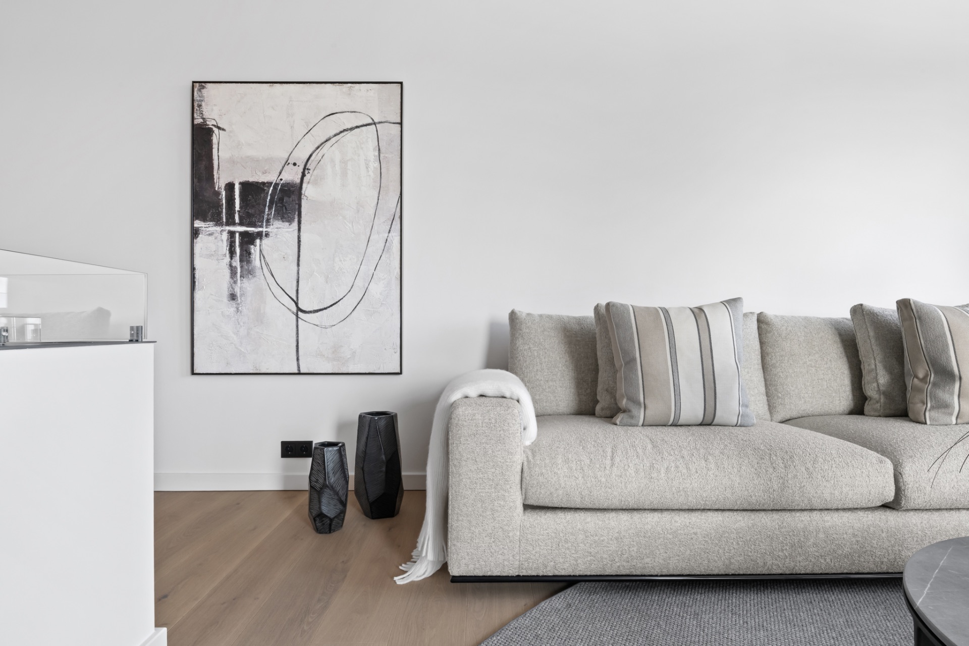 Helles Wohnzimmer mit beigem Sofa, abstraktem Bild an der Wand und zwei dunklen Vasen auf Holzboden.