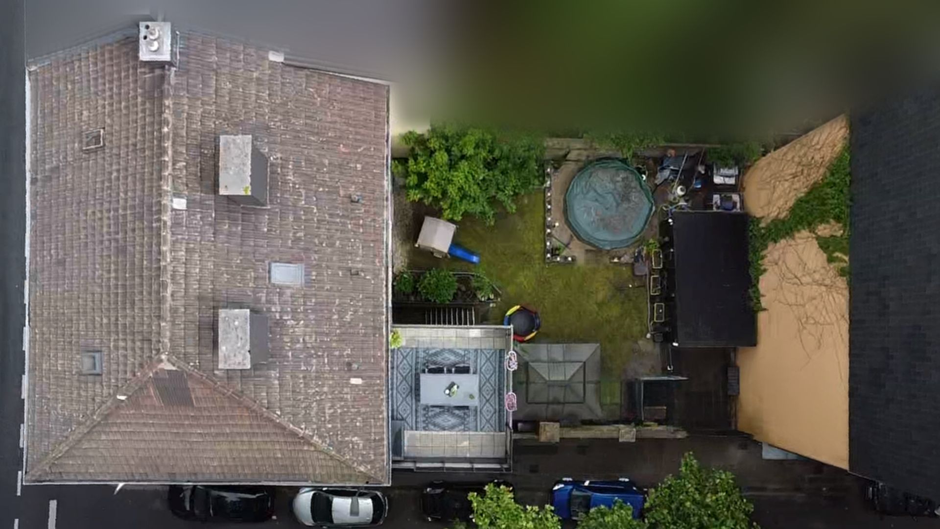 Luftaufnahme eines Hauses mit Garten, Trampolin und mehreren Autos am Straßenrand.